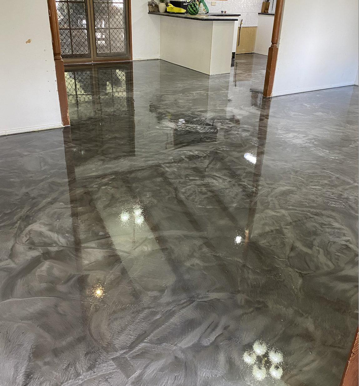 Indoor space with a highly reflective, wet concrete floor, showing reflections of the ceiling lights and surrounding doors and furniture.