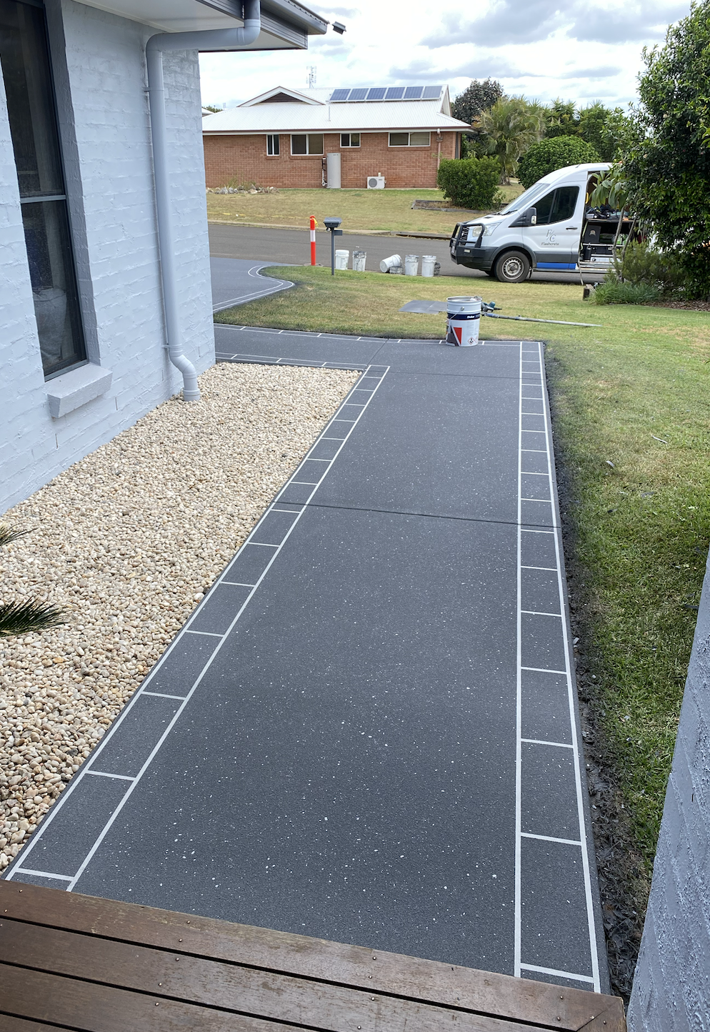 A newly paved sidewalk next to a house with a white brick wall, a window, and a gravel bed. In the background, a lawn, a street with a van parked, and another house with solar panels on the roof are visible.
