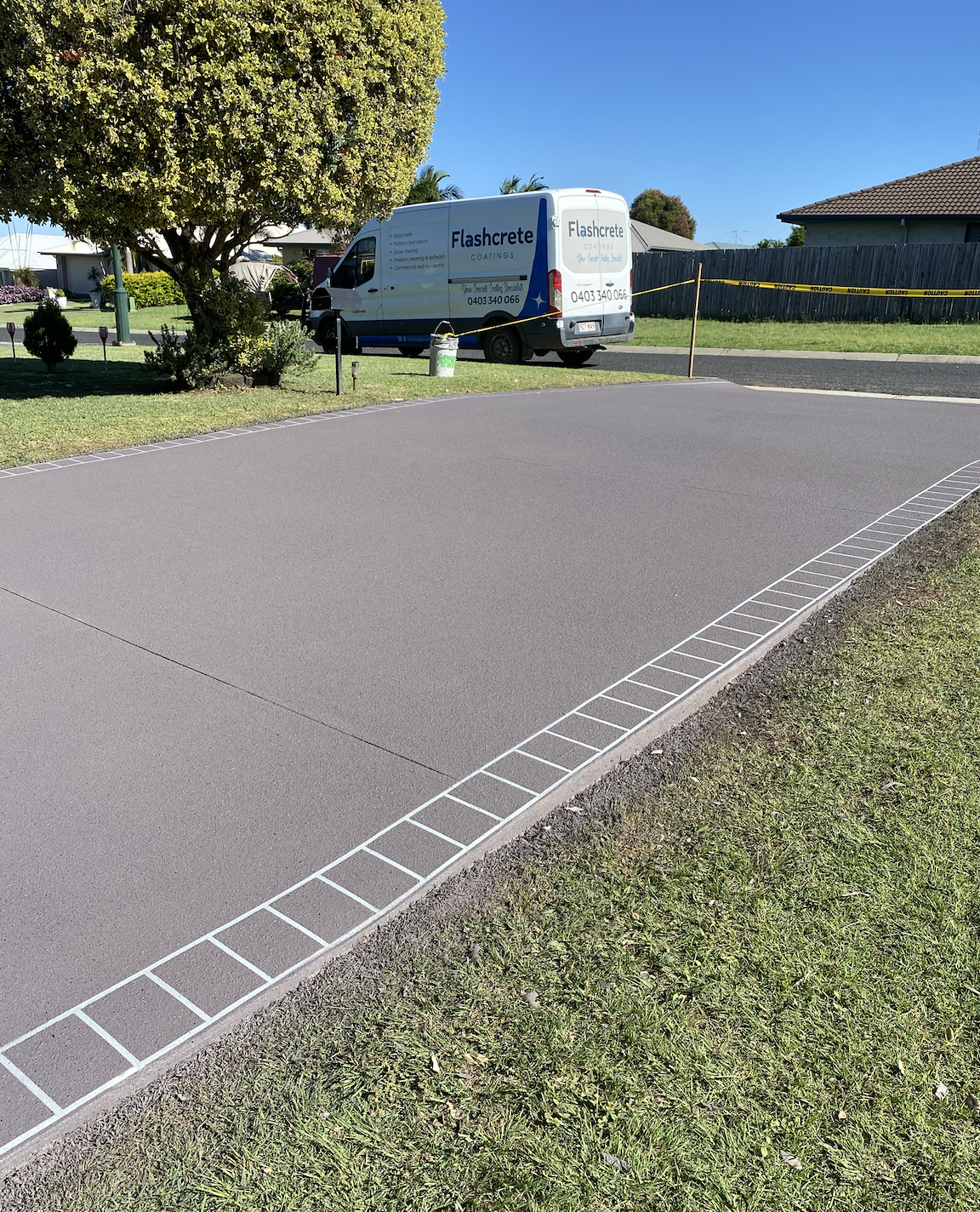 A residential driveway with a newly paved surface, bordered by decorative bricks. A white van with advertising for Flashcrete Coatings is parked in the background near a large tree, with yellow caution tape around the area.