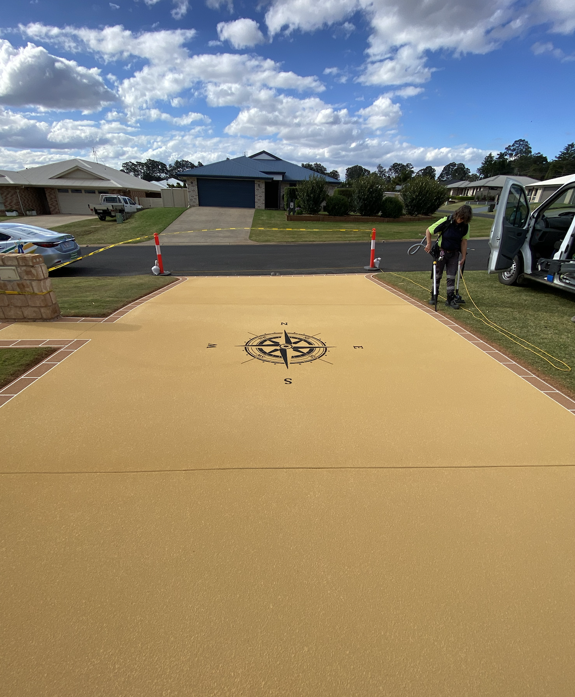 A newly paved driveway with a decorative compass rose design in the center, being prepared or serviced by a worker near a white van in a suburban neighborhood on a partly cloudy day.