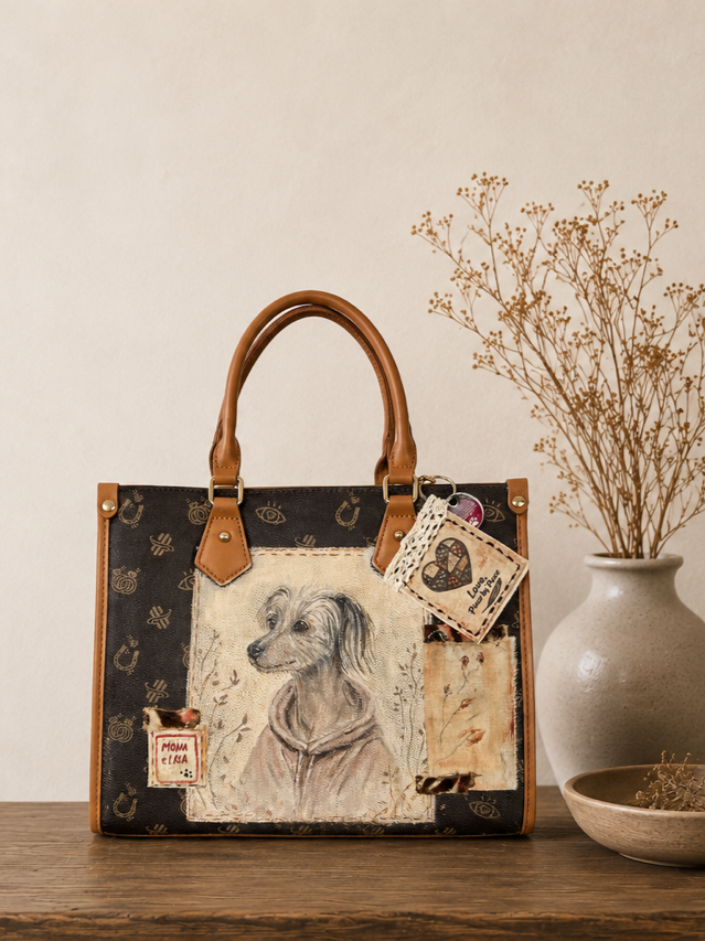 A black handbag with tan handles and accents, decorated with various patches and artwork, including a dog wearing a hoodie, placed on a wooden surface next to a vase with dried flowers.
