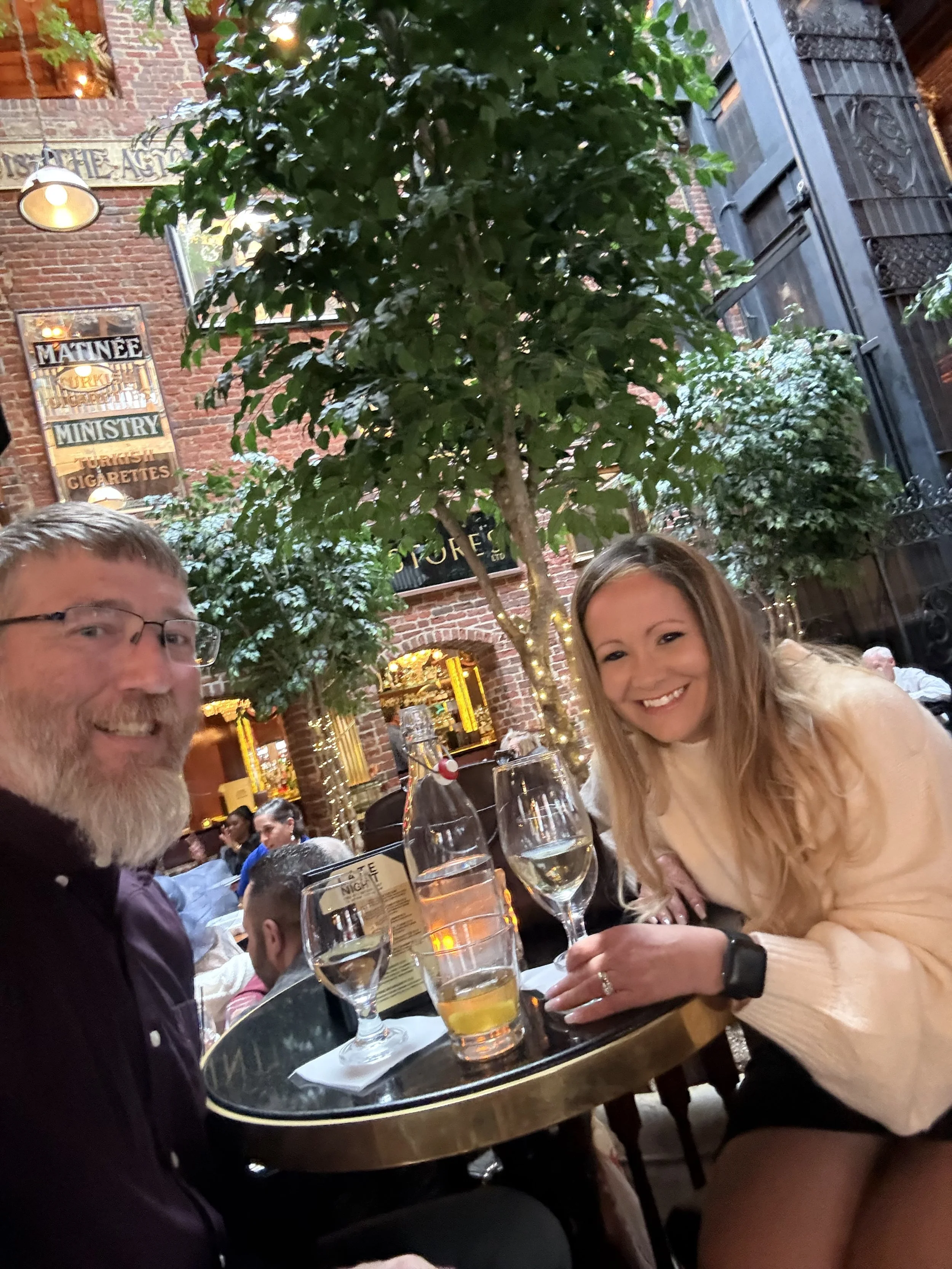 Two people seated at a table in a restaurant with brick walls, green plants, and decorative lighting; they are smiling and have drinks in front of them.