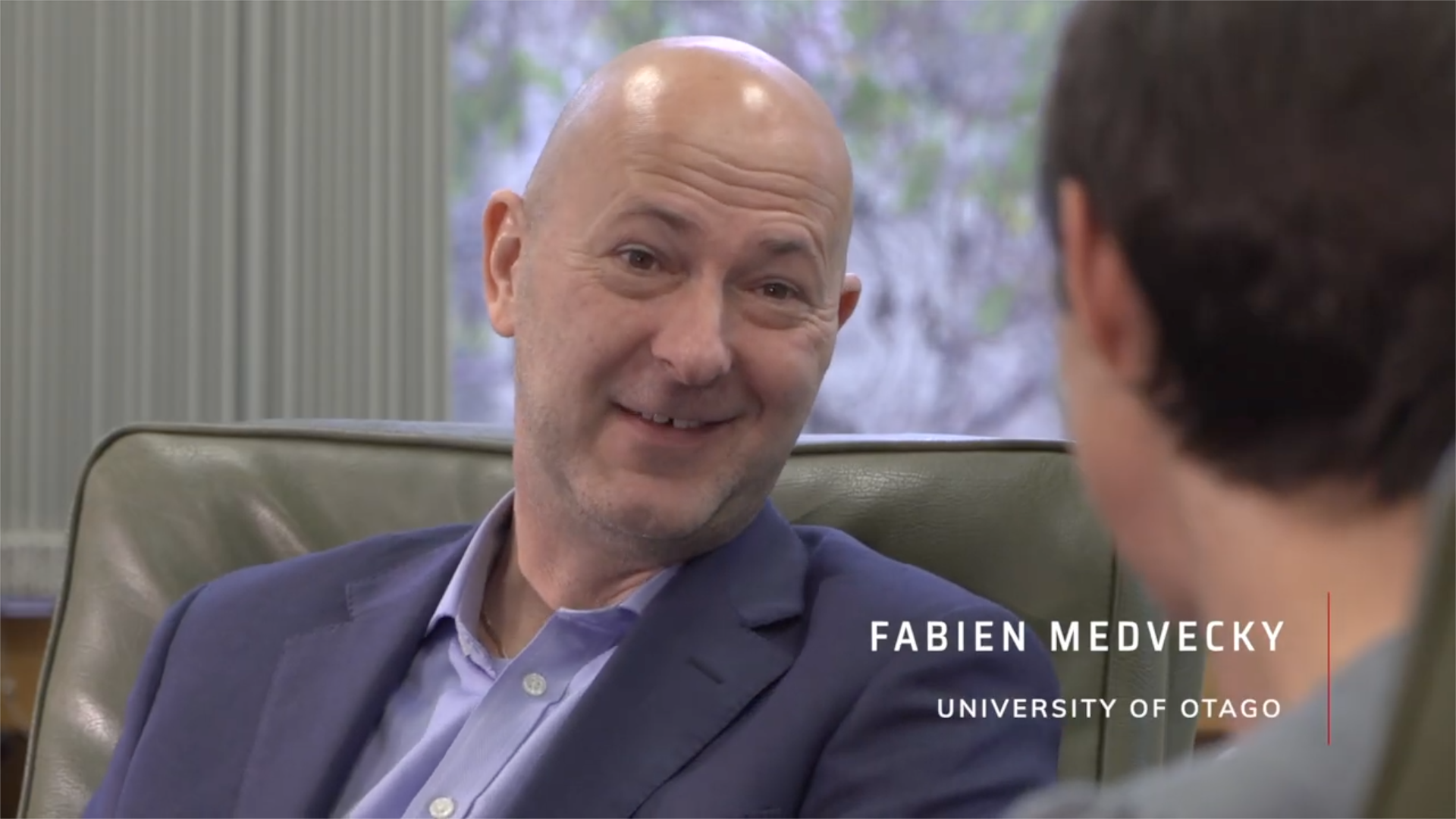 A man with a bald head and wearing a dark suit is smiling and talking to someone. The background shows a window with blurred outdoor scenery. The partial text on the screen reads 'FABIEN MEDVECKY' and 'UNIVERSITY OF OTAGO.'