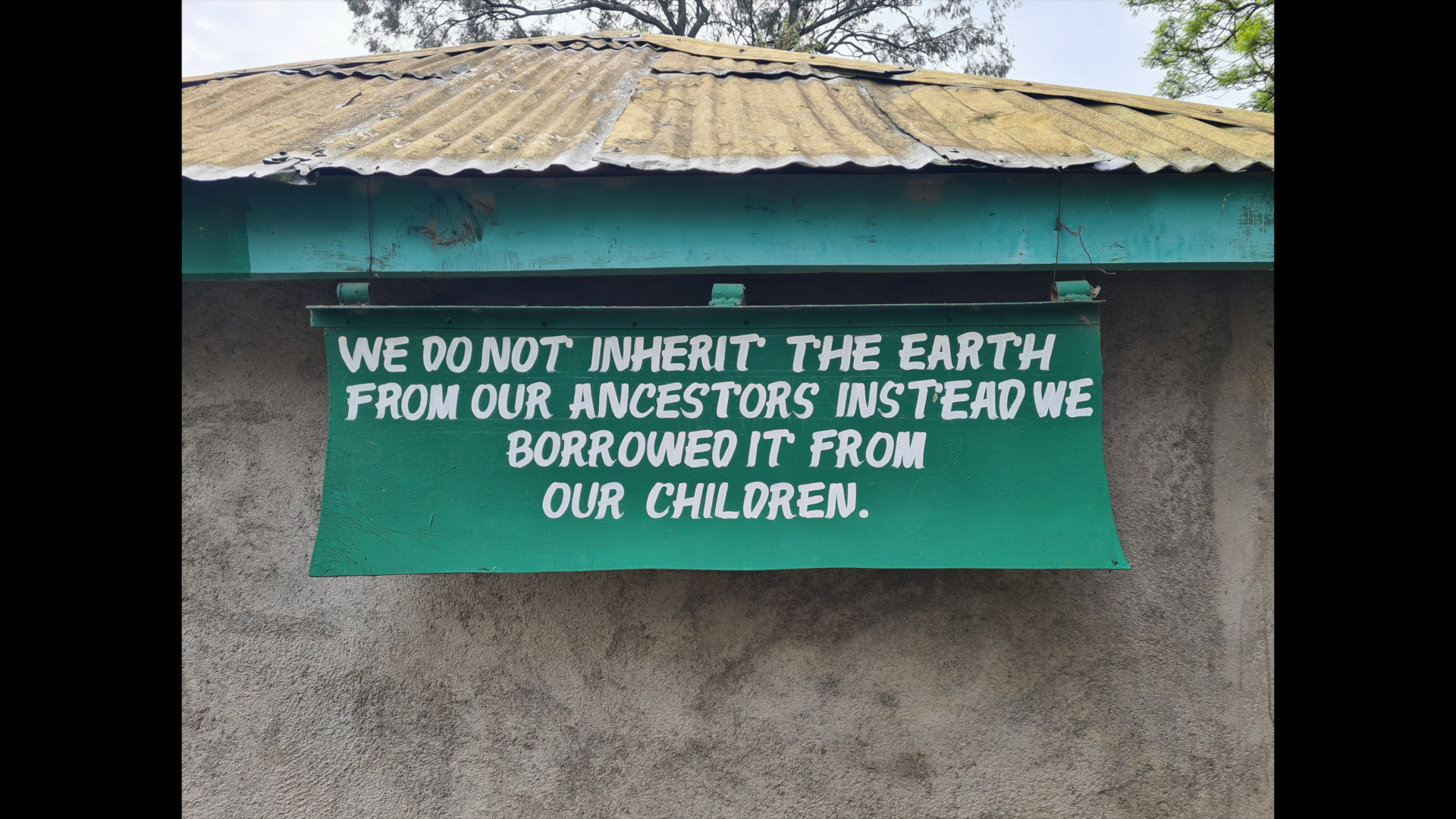A green sign with white text hanging on a wall that reads, "We do not inherit the earth from our ancestors instead we borrowed it from our children." The wall is textured, and the roof above appears old and weathered with moss or dirt.