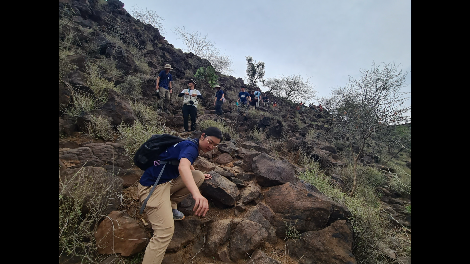 Group of hikers climbing a rocky slope on a mountain trail, some using hands for balance, surrounded by sparse desert vegetation and leafless trees.