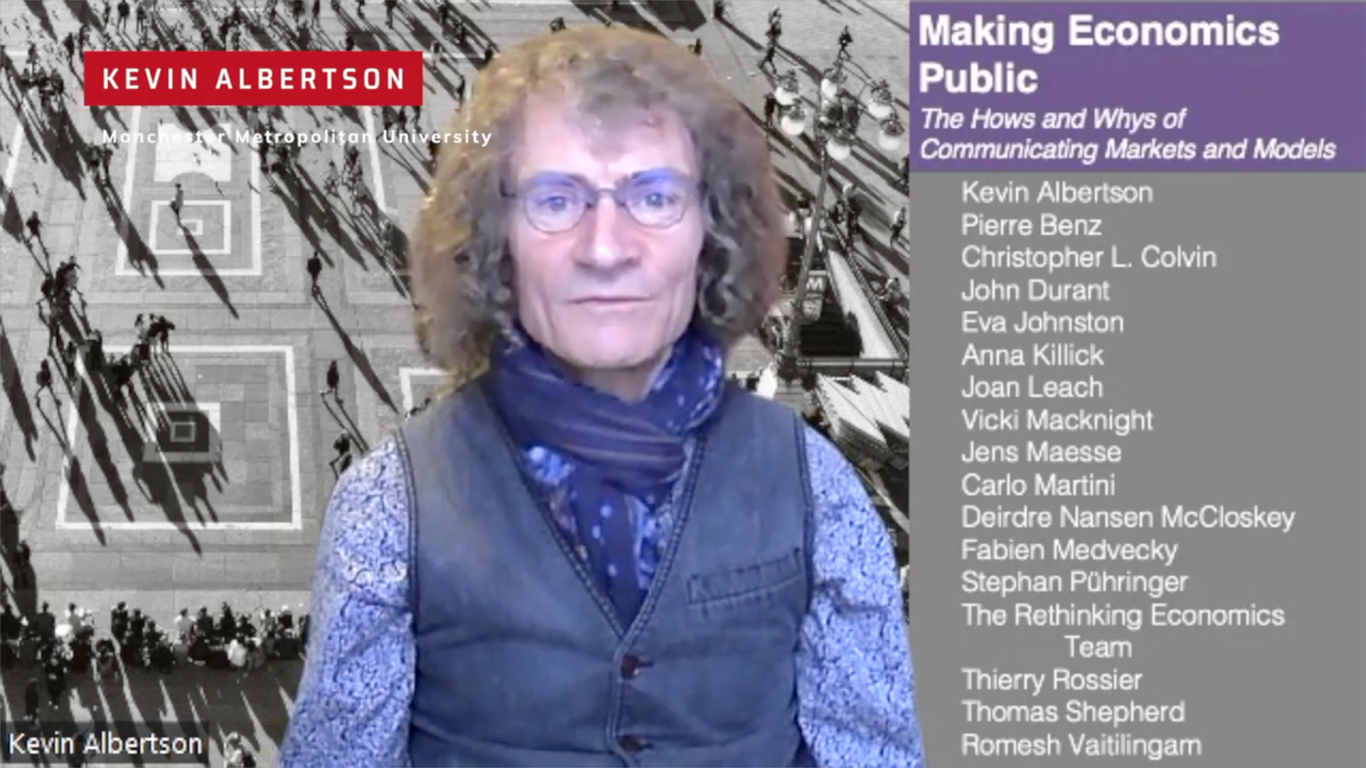 A man with curly hair and glasses wearing a blue scarf, standing in front of a background showing a busy outdoor scene and a panel of text listing names and titles related to economics.