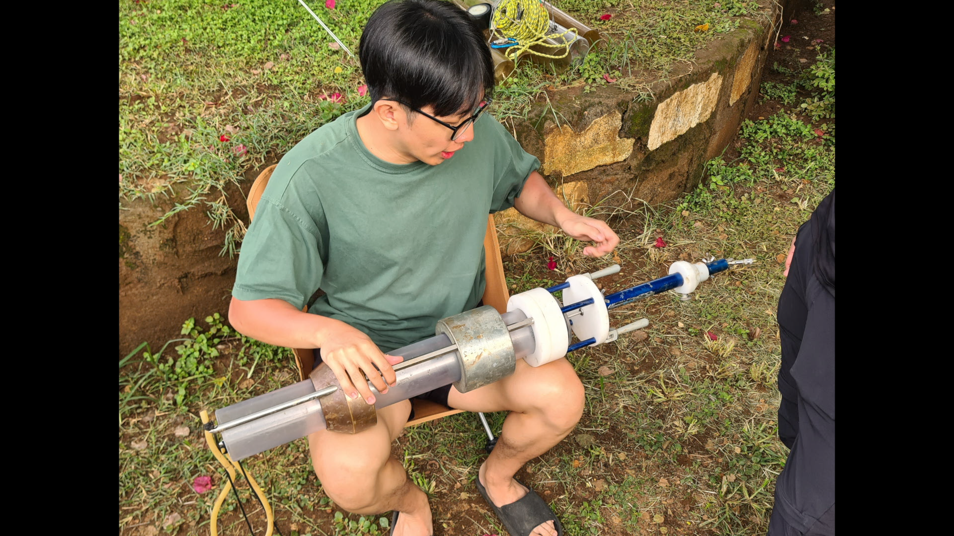 A man sitting outdoors on a chair, holding a large scientific instrument resembling a telescopic device, with a garden background, wearing a green T-shirt, glasses, and sandals.