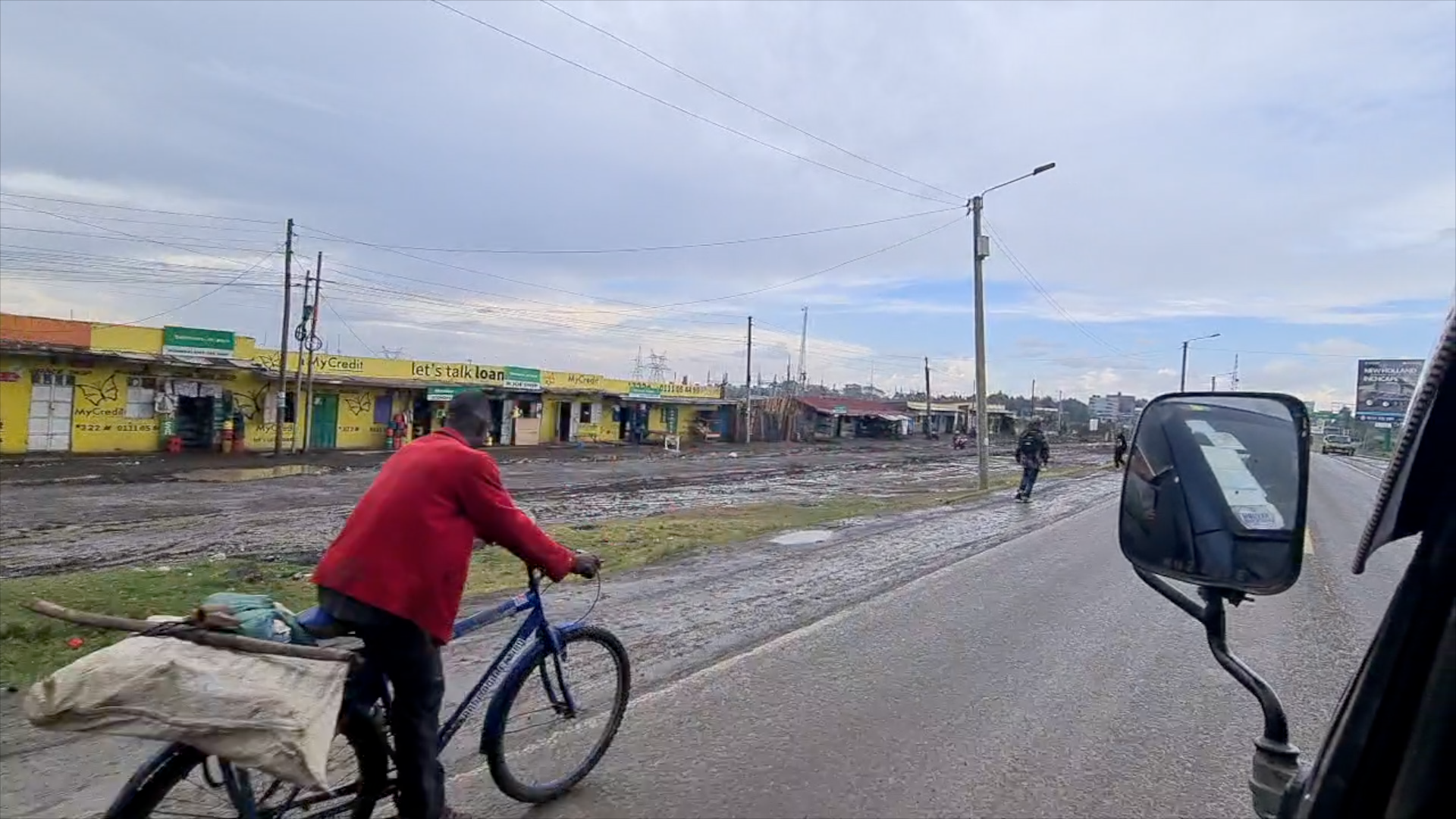 A street scene with a bicycle rider wearing a red jacket, a person walking, and buildings with yellow walls advertising loans, under a cloudy sky, water puddles and mud on the road, and utility poles and wires.