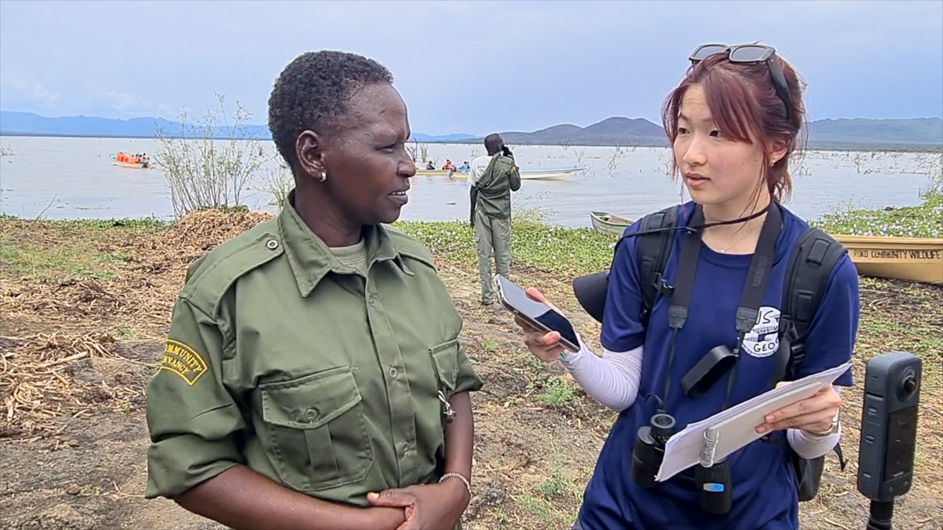 A woman in a blue shirt and glasses on her head is interviewing a woman in a green uniform near a river, with boats and people in the background.