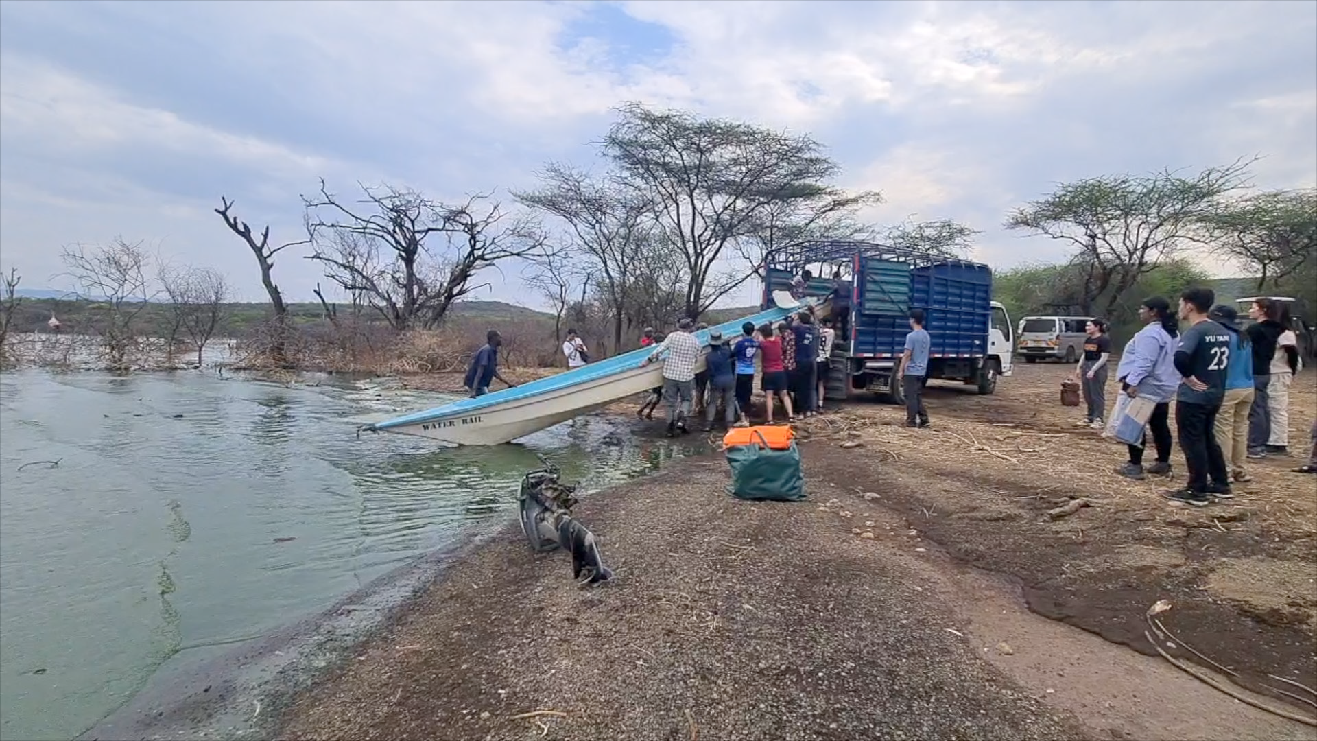 A group of people is launching a boat into a body of water from a rocky shoreline with a truck. The scene is outdoors with sparse trees and cloudy sky.