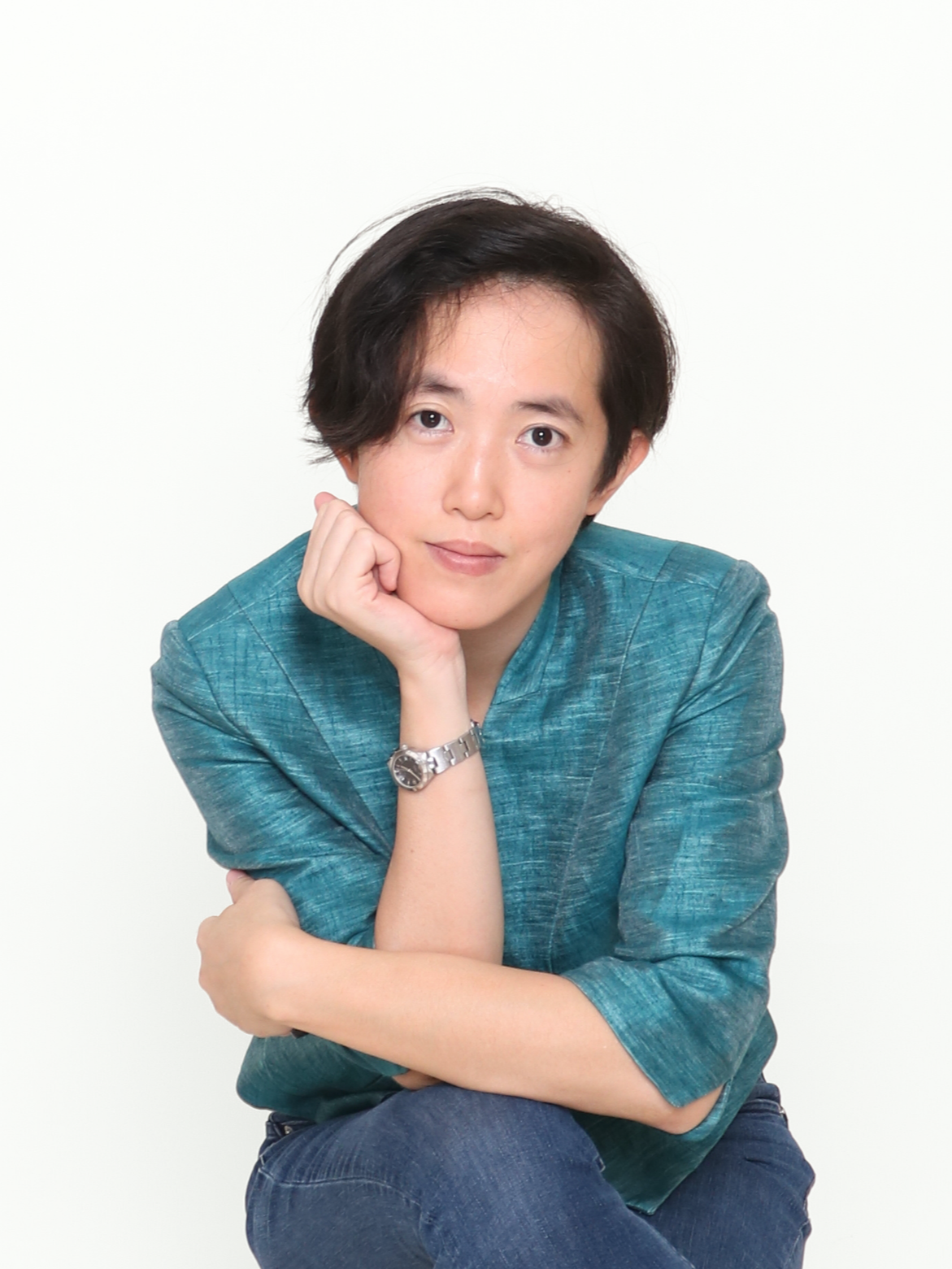 Portrait of a woman with short dark hair wearing a teal top and jeans, sitting with her left elbow on her knee, resting her chin on her hand, against a plain white background.
