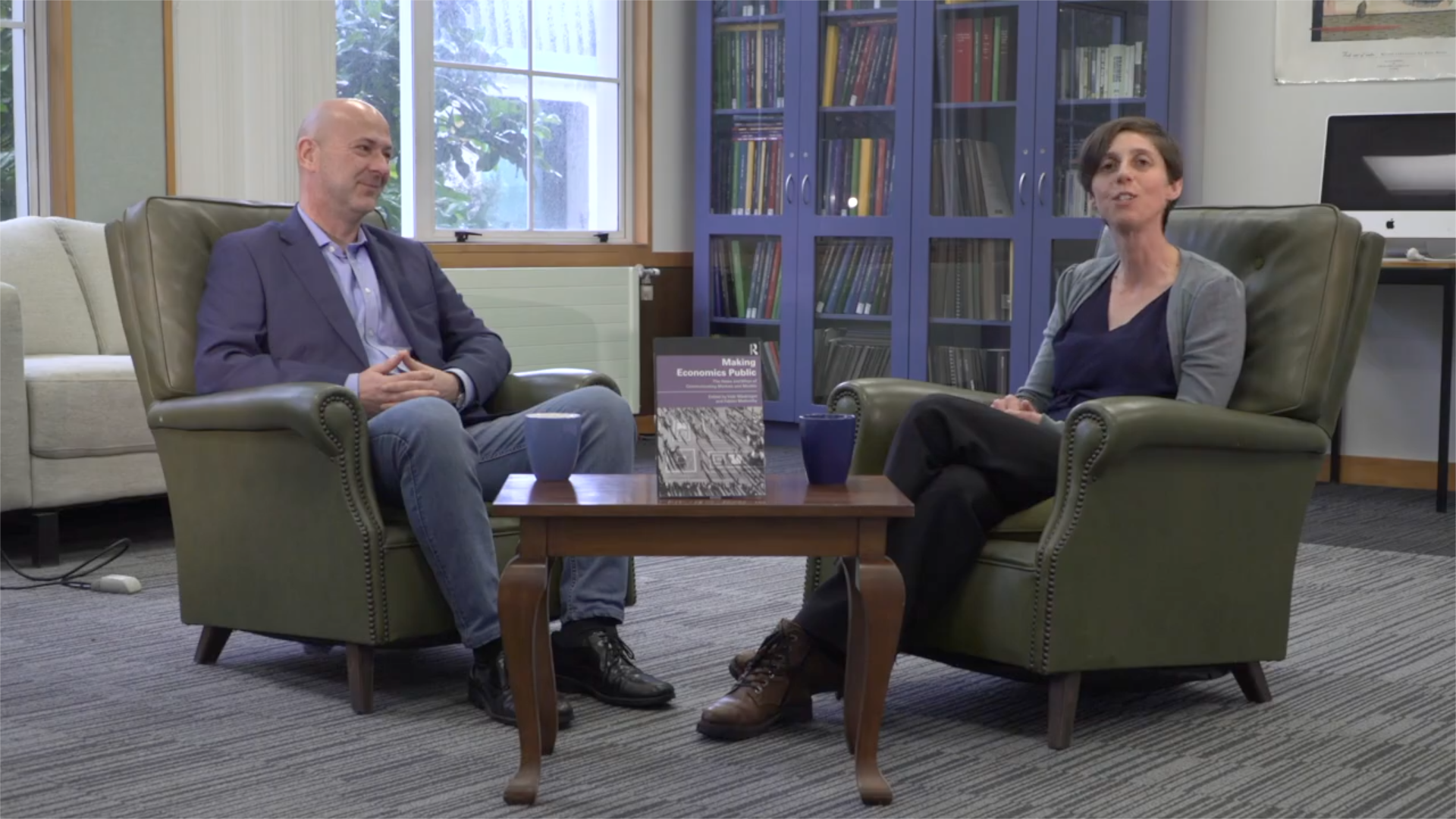 Two people, a man and a woman, sitting in green armchairs in a library or office, engaged in conversation. A small wooden table with a book titled 'Making Economics Public' and two blue mugs is between them. Behind them are bookshelves filled with colorful books, a window, and a computer on a desk.