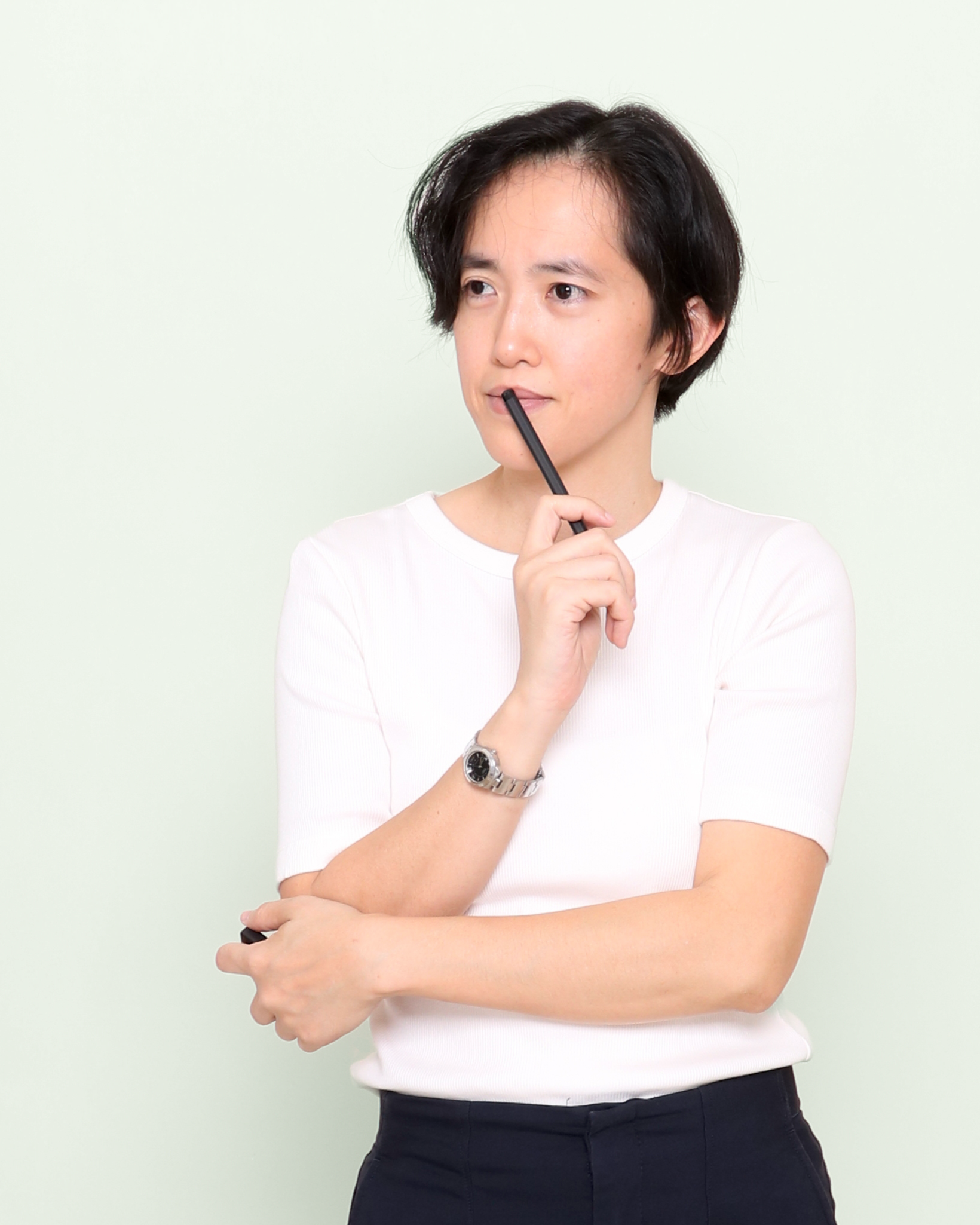 A woman with short black hair, wearing a white top and black pants, standing against a light-colored background, holding a pen near her lips and looking thoughtfully.