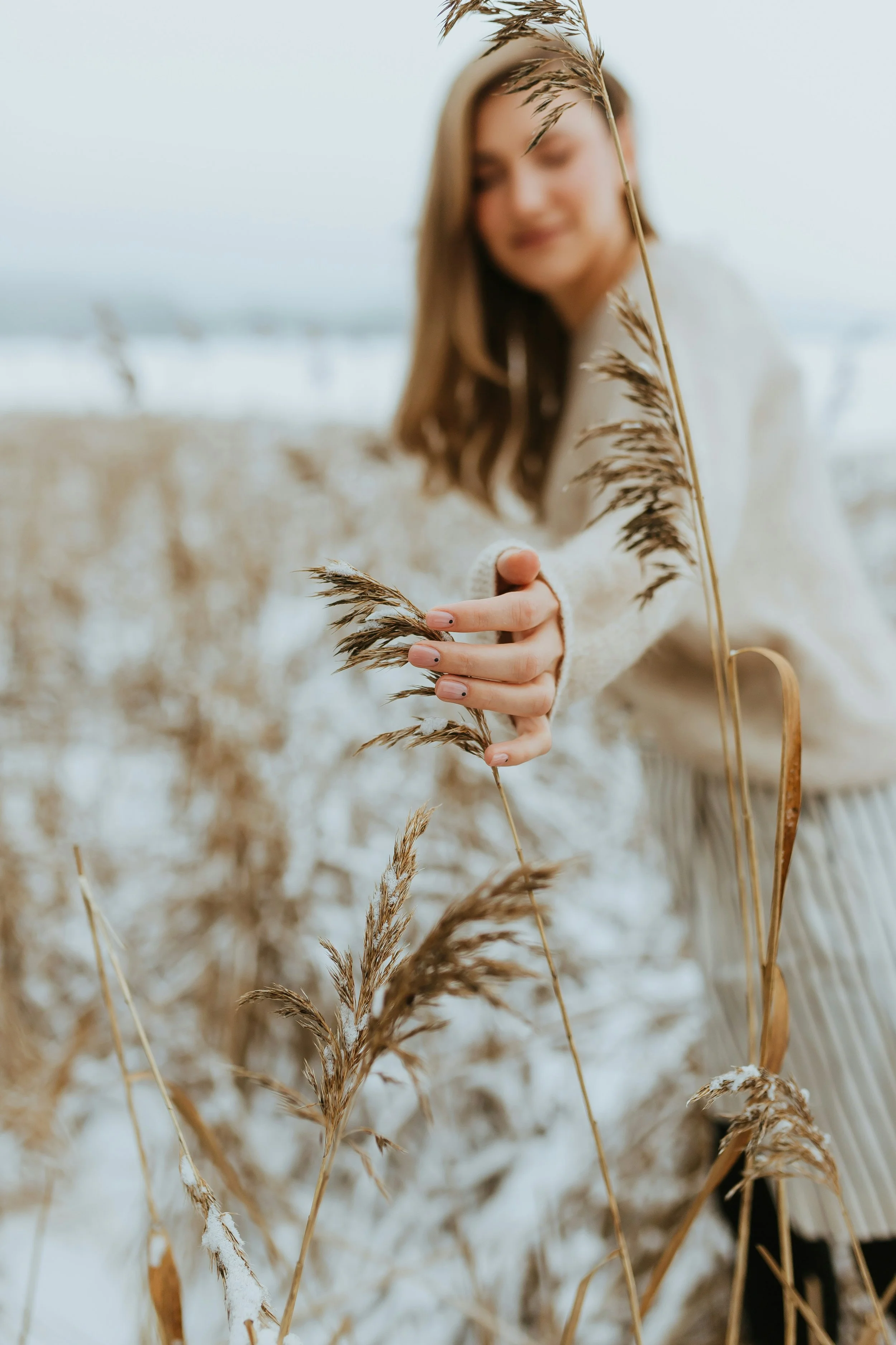 A woman in a white sweater holds a tall dried grass stalk in a field of similar grasses during daytime with an overcast sky.