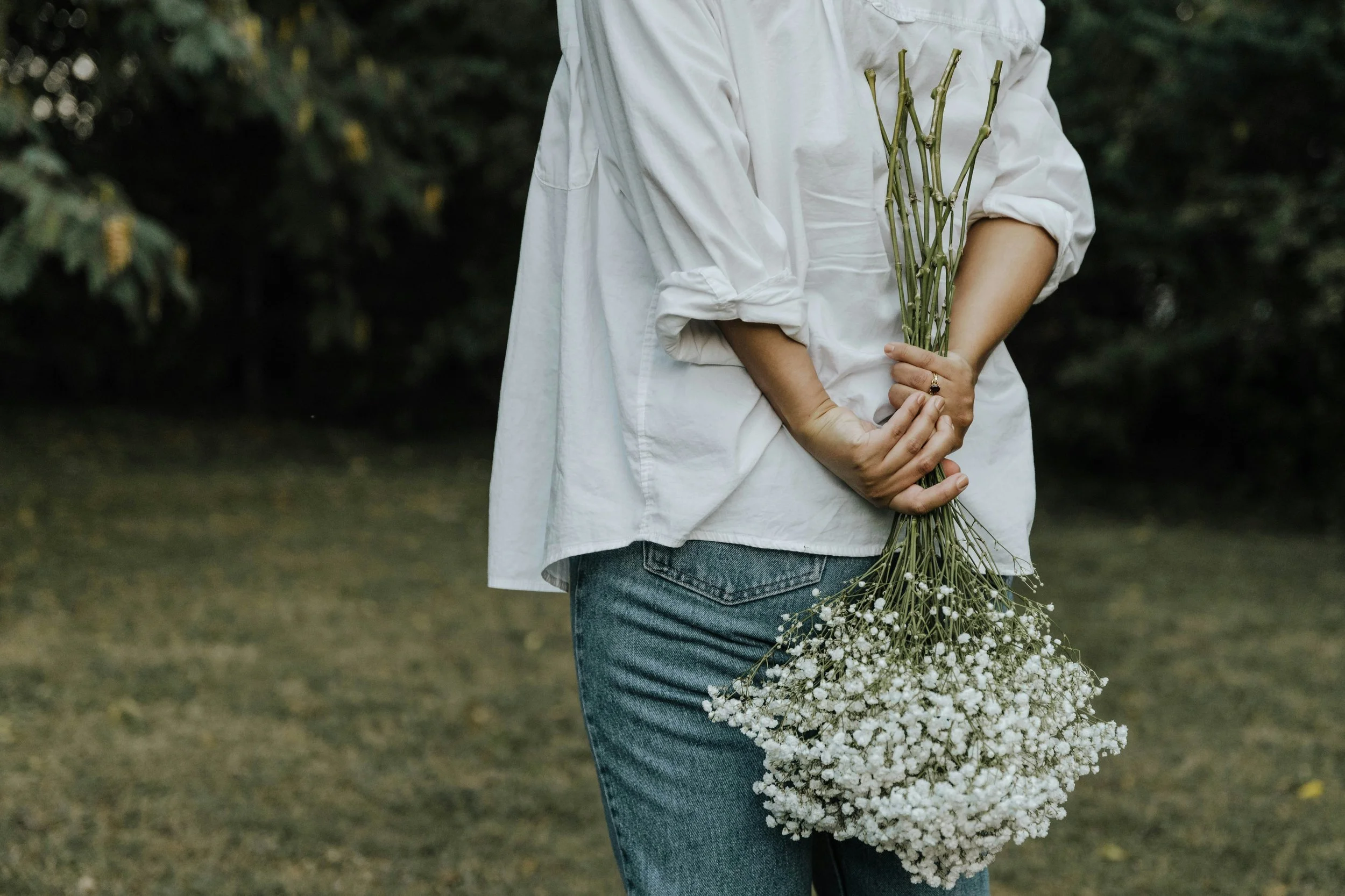 Person in white shirt holding a bouquet of small white flowers in an outdoor setting with greenery in the background.