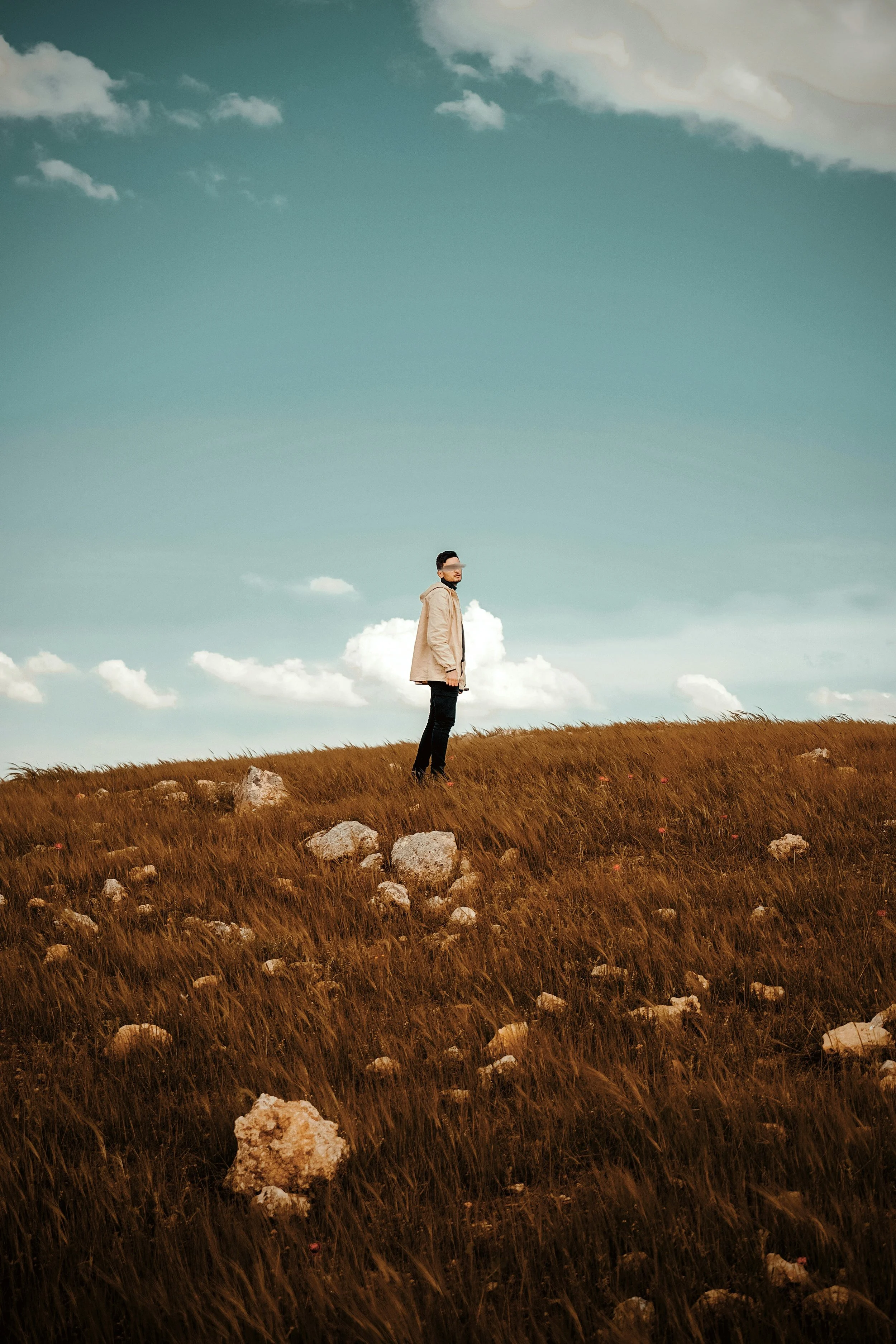 A man standing in a grassy field with rocks, wearing a beige jacket and sunglasses, under a cloudy sky.