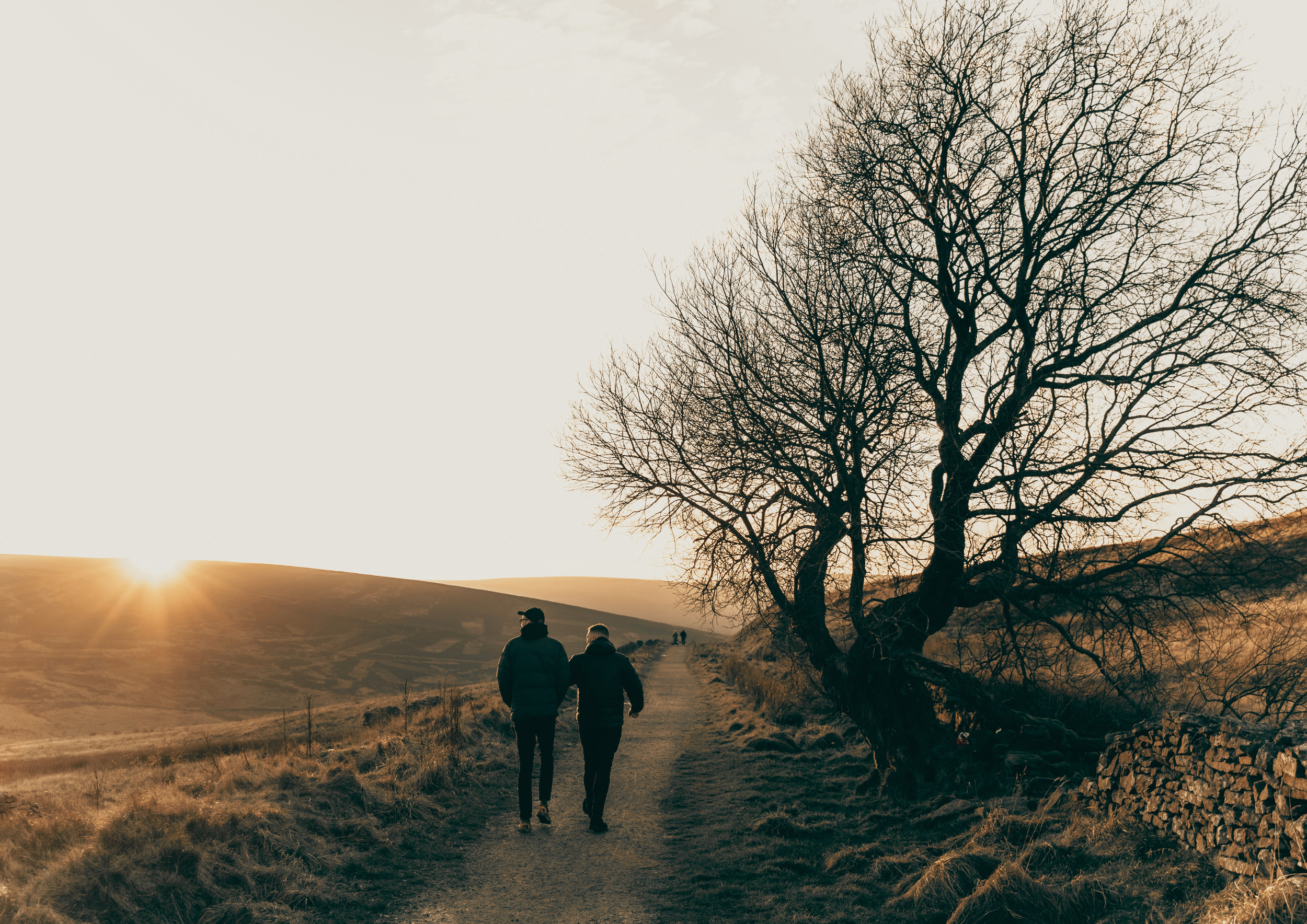 Two people walking on a dirt trail in a hilly landscape at sunset, with barren trees and rolling hills in the background.