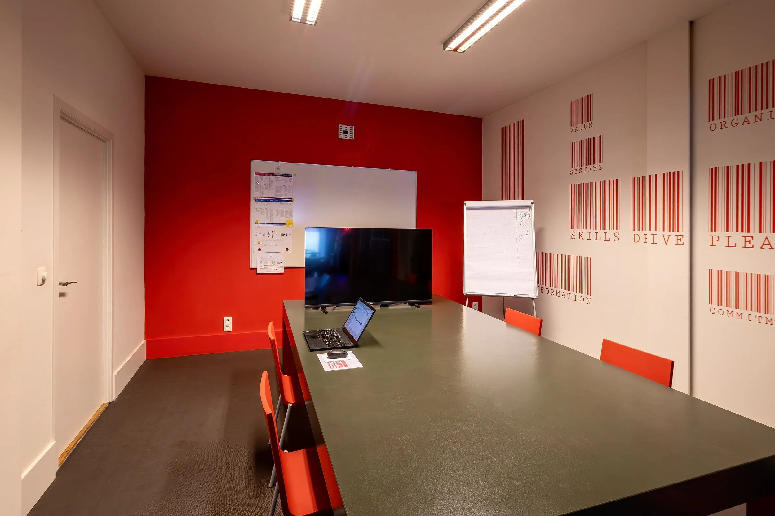 A conference room with a large green table, red chairs, a whiteboard, a TV, a laptop, and a flip chart. The wall has a red accent color with text and barcode graphics.