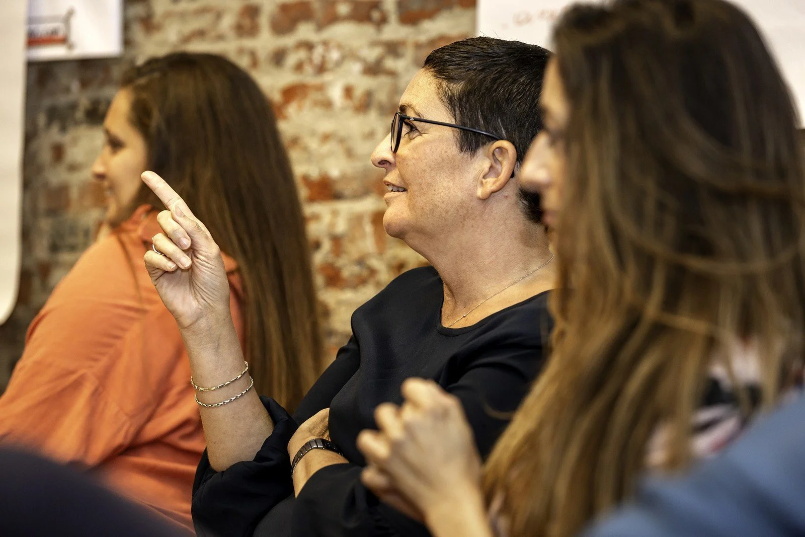 A group of people sitting in a row, with the focus on a woman with short dark hair, glasses, and a black top, smiling and pointing, against a brick wall background.