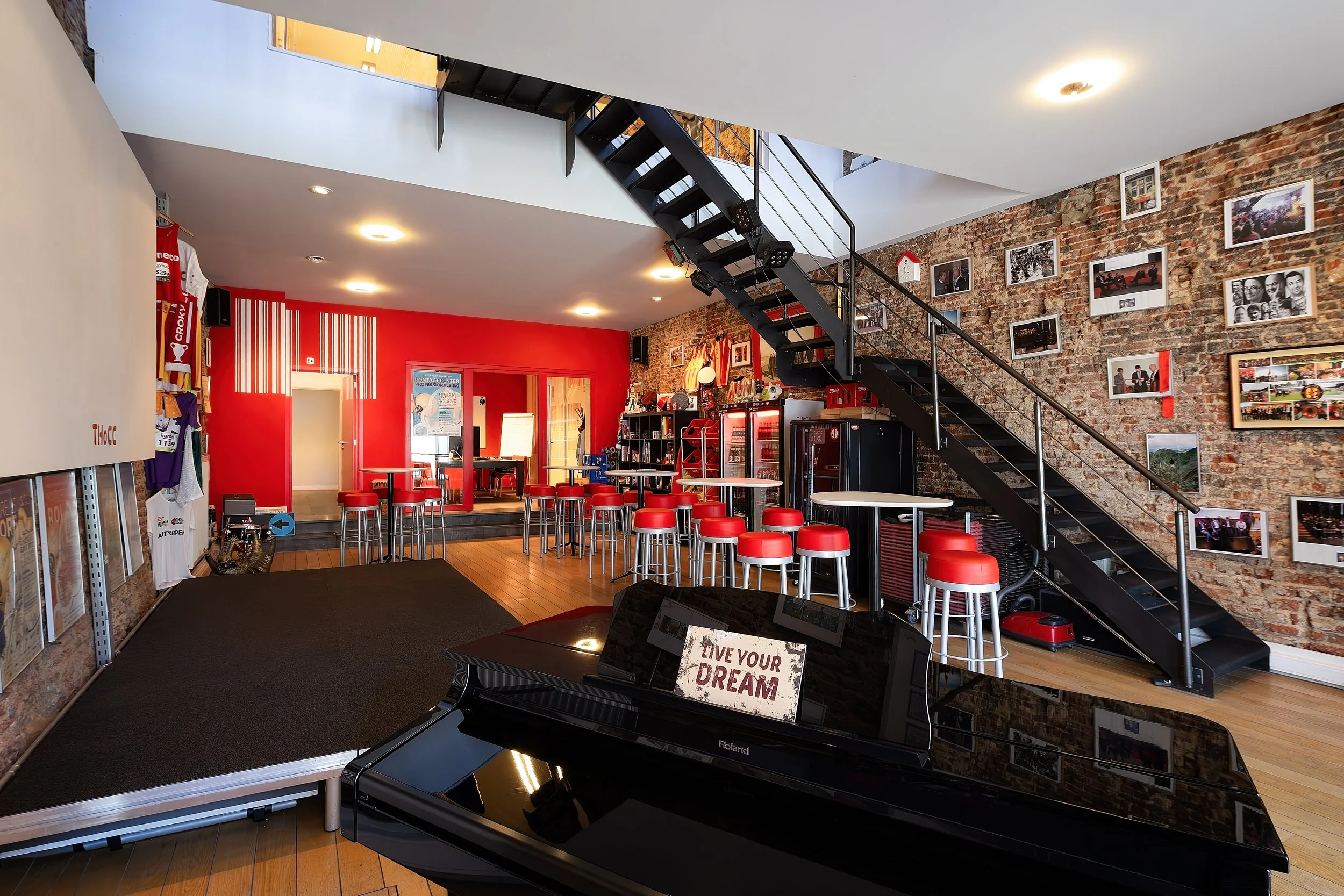 Interior of a cafe or lounge area with a black grand piano in the foreground, decorated with a sign that says "Live Your Dream." The space features exposed brick walls with framed photos, red and black furniture, and a staircase with black steps and metal railing leading to an upper level.