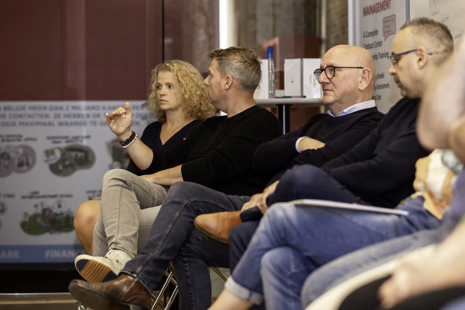 A group of people sitting in a panel discussion or conference, with some listening attentively and one man in black raising his hand.