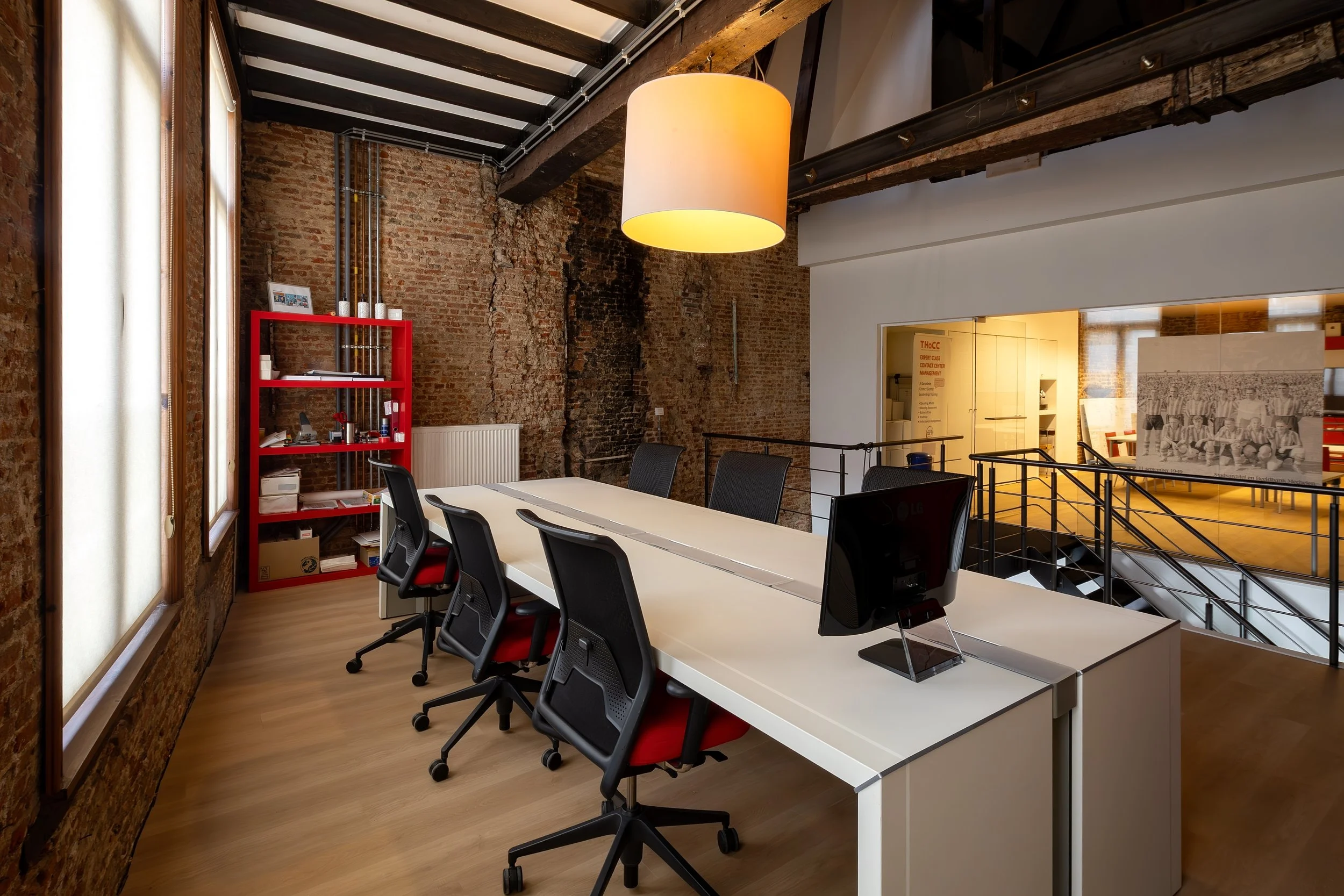 A modern industrial-style office space with exposed brick walls, wooden beams, and large windows with white shades. There is a long white table with black office chairs, a red shelving unit with documents and supplies, and a yellow calendar or poster on the wall behind. A large hanging lamp provides lighting, and there is a black railing surrounding a staircase or balcony area.