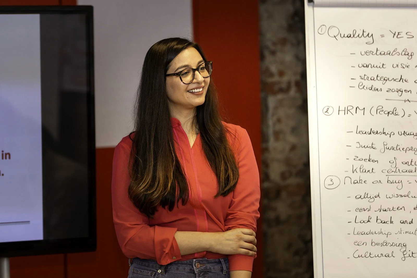 A woman with long dark hair and glasses wearing a bright red shirt, standing in front of a whiteboard with handwritten notes in a room with brick and red walls.