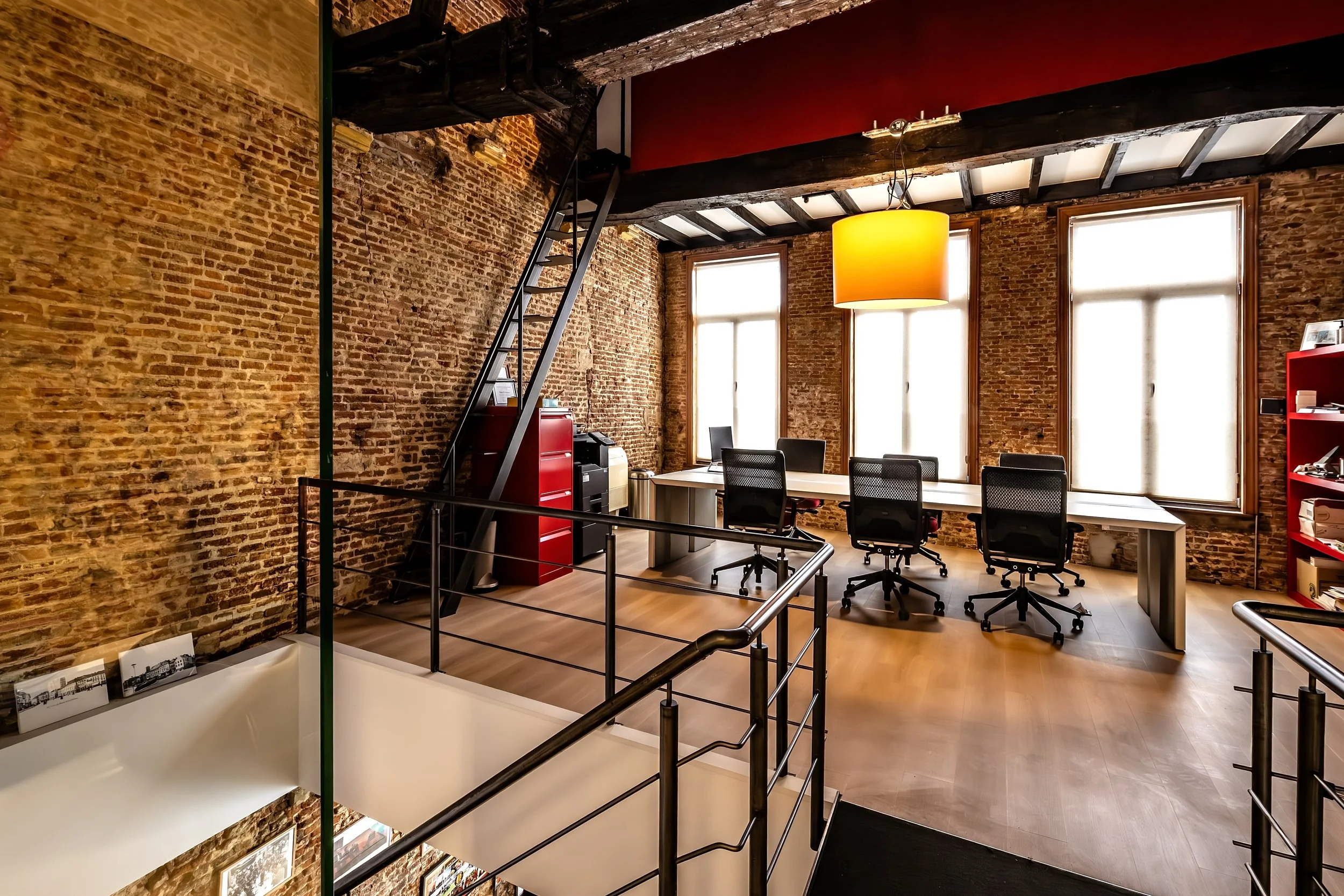 An office space with exposed brick walls, large windows, and modern furniture. There are black chairs, a white table, and a yellow pendant light hanging from the ceiling. A staircase and some framed photographs are visible in the foreground.