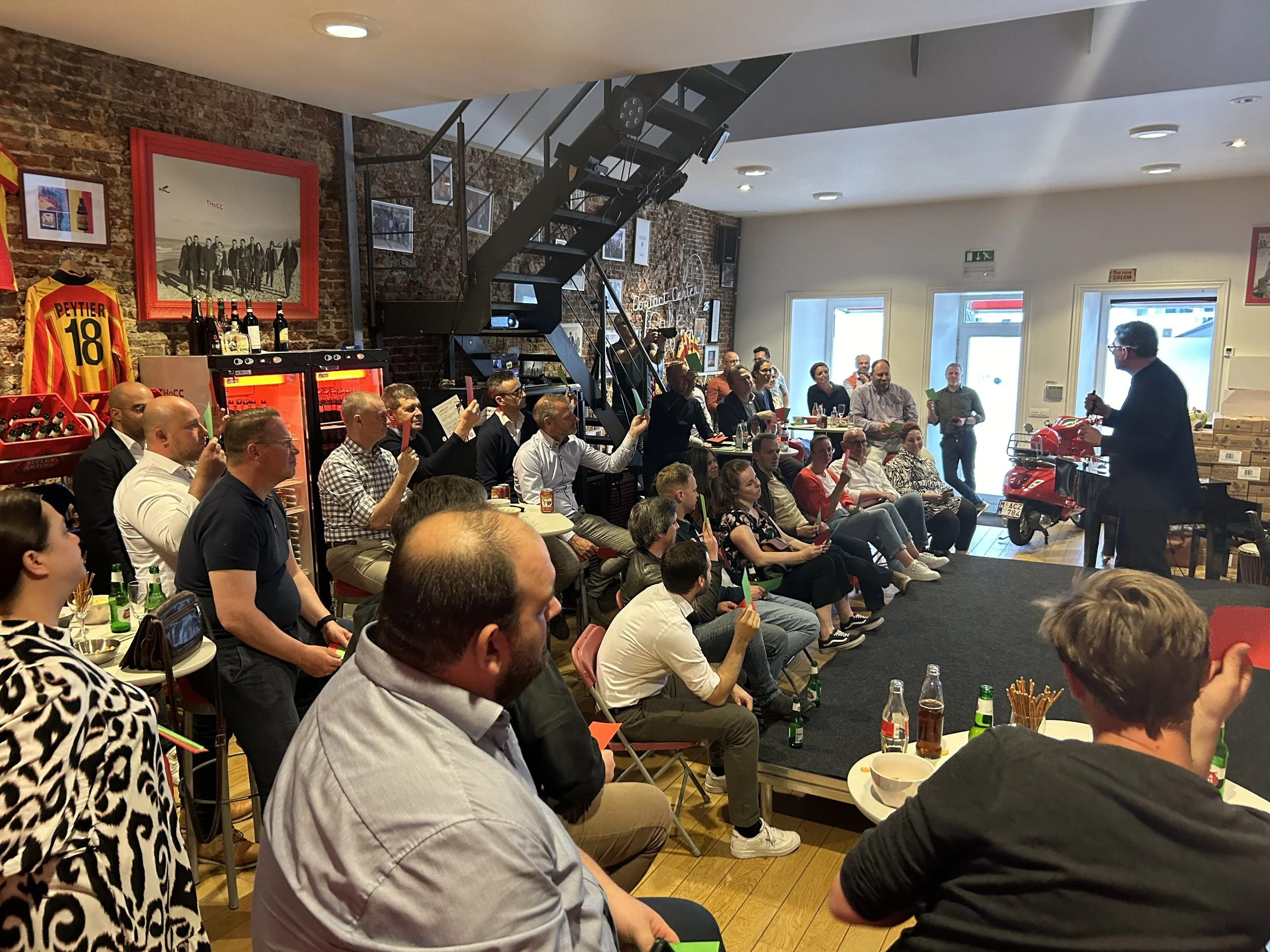 An indoor gathering with people seated and standing, listening to a man giving a speech. There are decorations and pictures on the brick wall, including a framed photo and sports jersey. Various drinks and snacks are on tables.