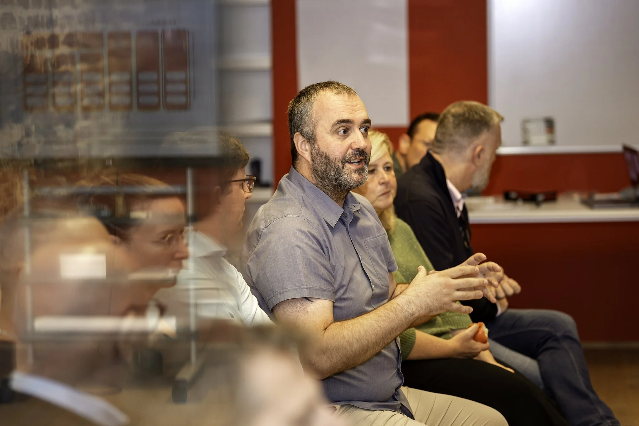 A group of people sitting at a meeting or conference, with one man in focus who has a beard and is wearing a gray shirt, listening attentively.