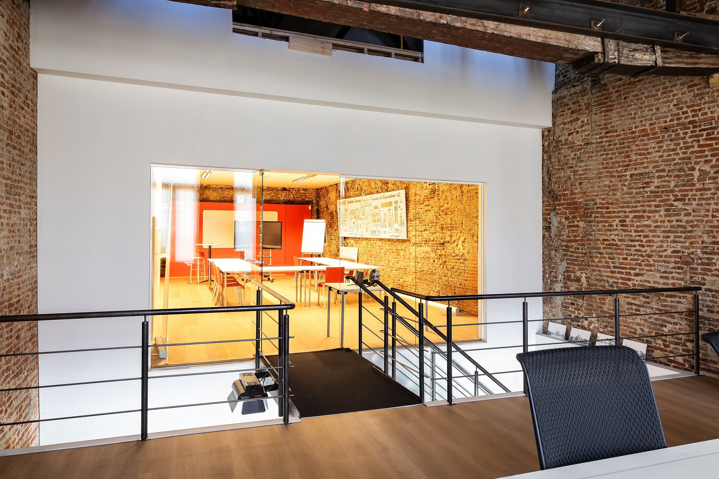 Modern office space with brick walls, glass windows, a black railing, and a conference room with orange walls and framed artwork.