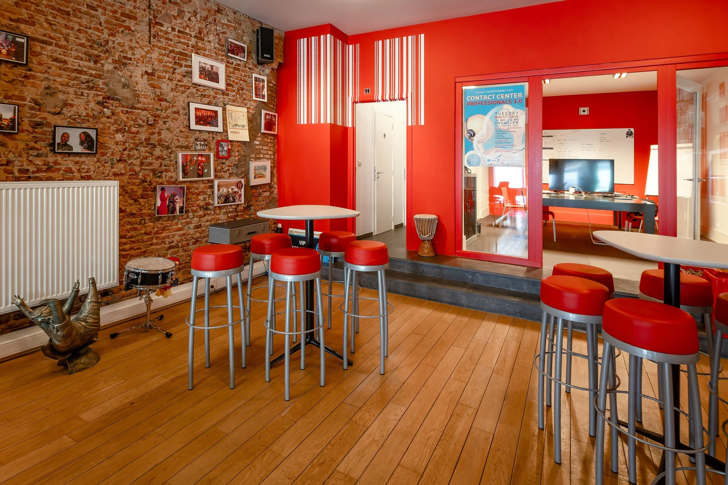 A modern interior space with a brick accent wall decorated with framed photos. There are high tables with red stools, a drum, and a decorative reindeer sculpture in the foreground. A bright red wall with striped lighting fixtures and an open doorway leading to an office area with a television and whiteboard are visible.