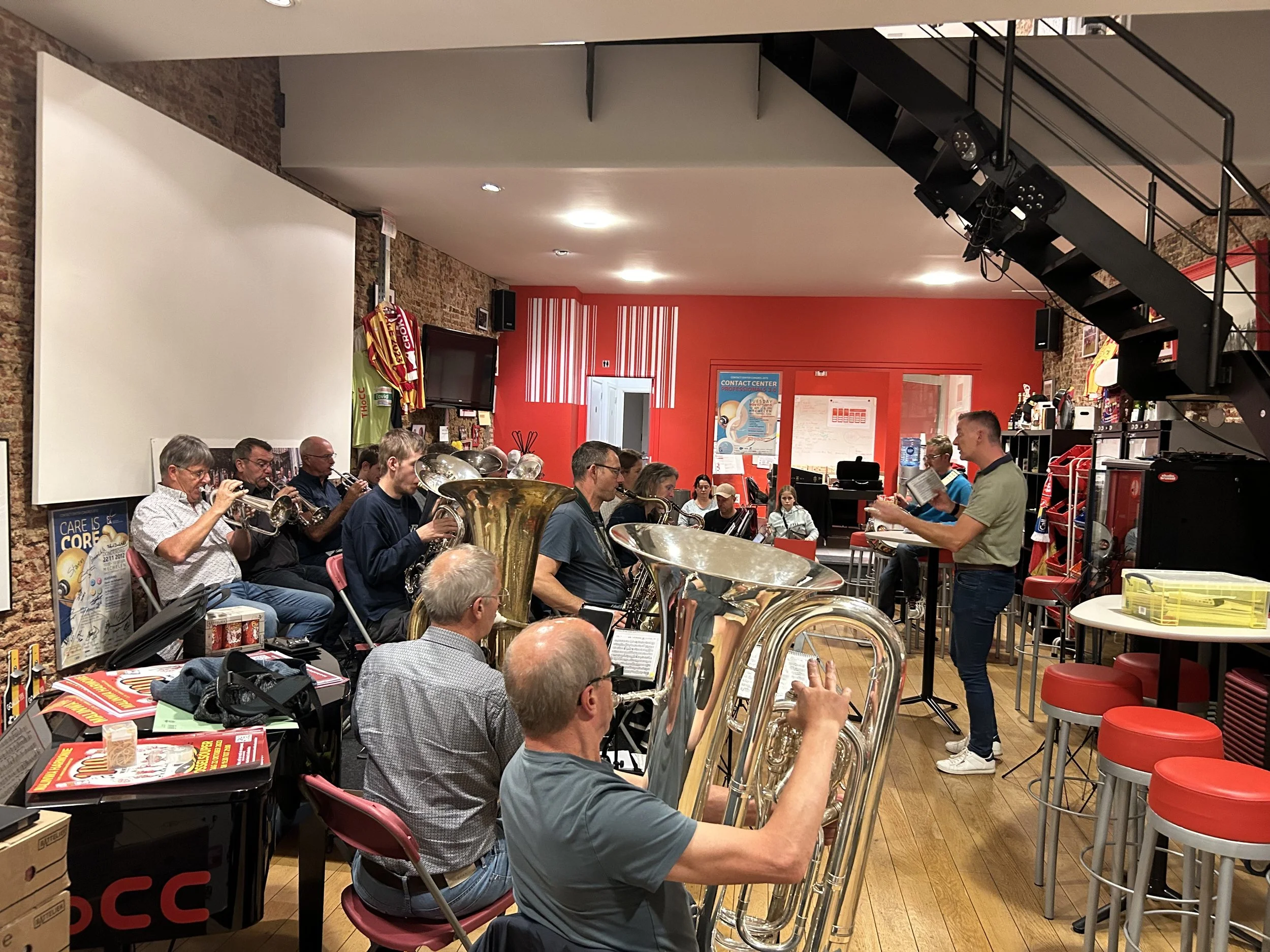Group of musicians playing brass instruments in a community center or music venue with red painted walls, tables, and people watching.