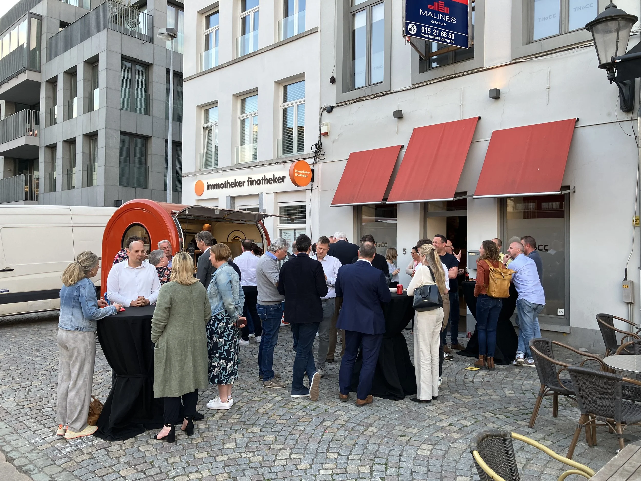 A group of people gathered outdoors on a cobblestone street in front of a building with .red and white awnings and a sign for Immotheker finotheker. Some people are talking and others are standing around tables.