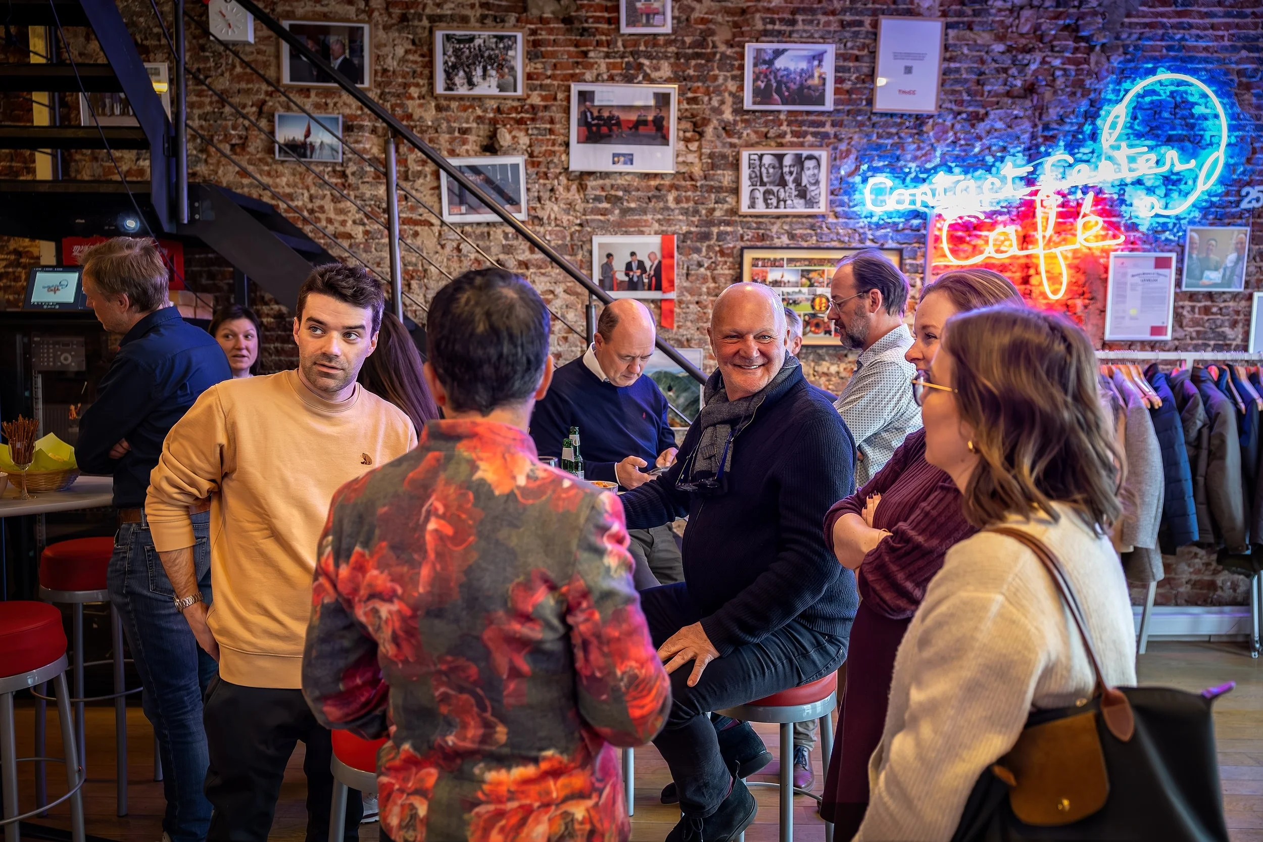 A group of people gathered in a restaurant or cafe with a brick wall decorated with framed photos and a neon sign that says 'Contact Center Cafe.' Some are seated and engaged in conversation, while others are standing nearby.