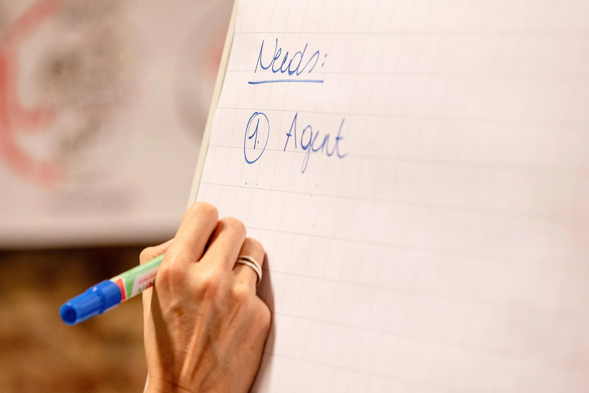 A person writing on a notepad with a blue and green marker, with notes that say 'Need:', 'P.:', and 'Augt' visible.