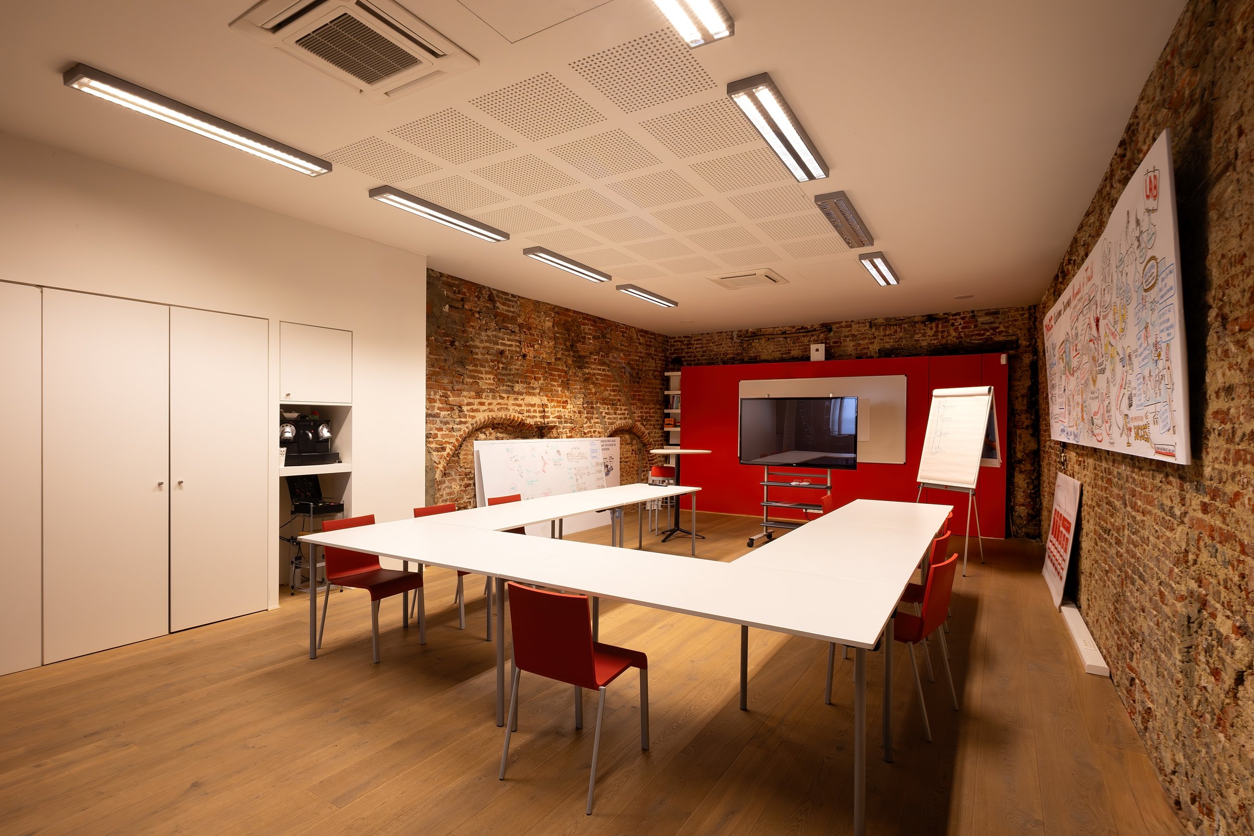 An office or conference room with brick walls, white tables arranged in a U-shape, red chairs, a large flat-screen TV, whiteboards, and a presentation poster on the wall. The ceiling has modern lighting fixtures.
