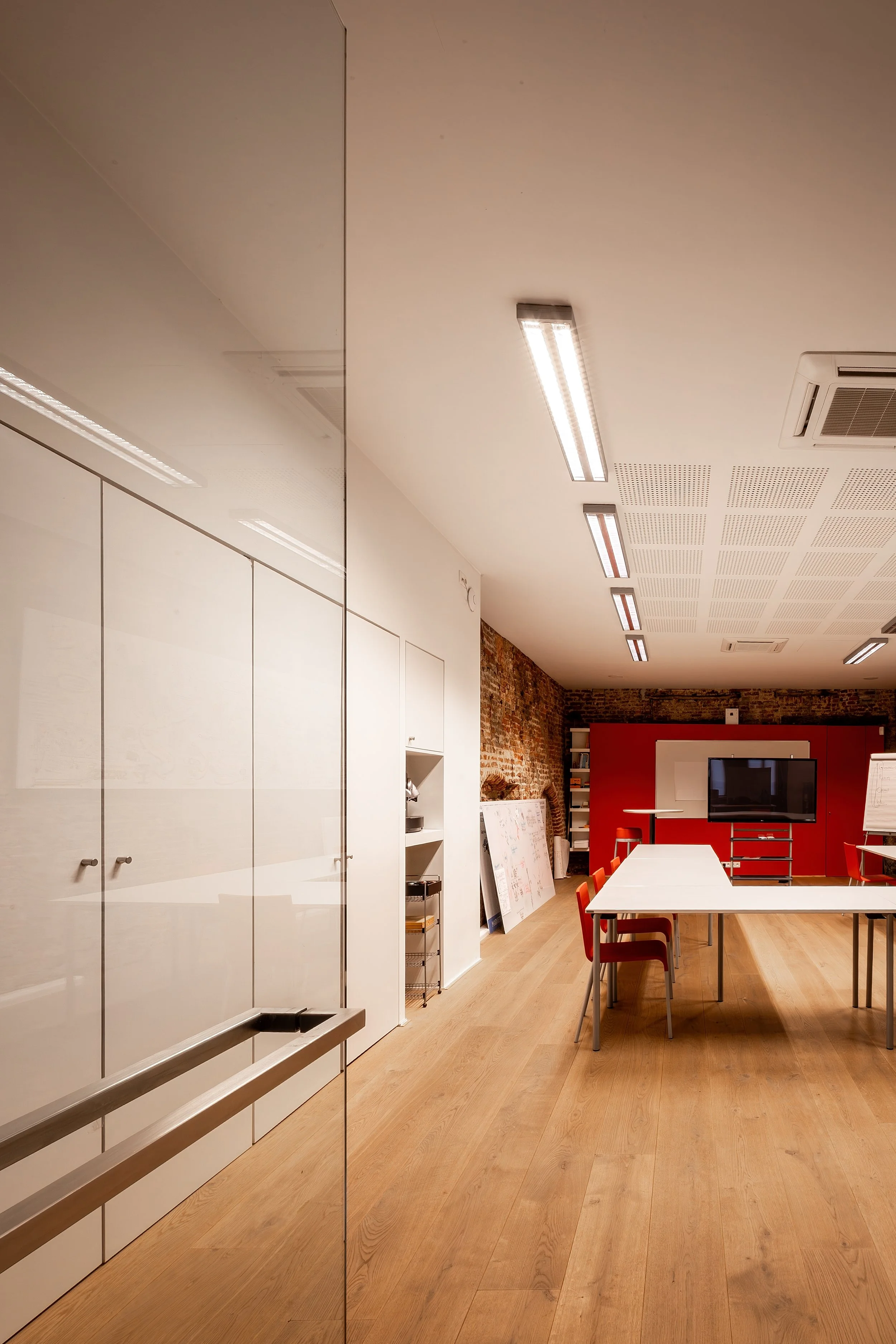 An office conference room with a large white table surrounded by red chairs, a brick wall, a red cabinet, a flat-screen TV, and whiteboards.