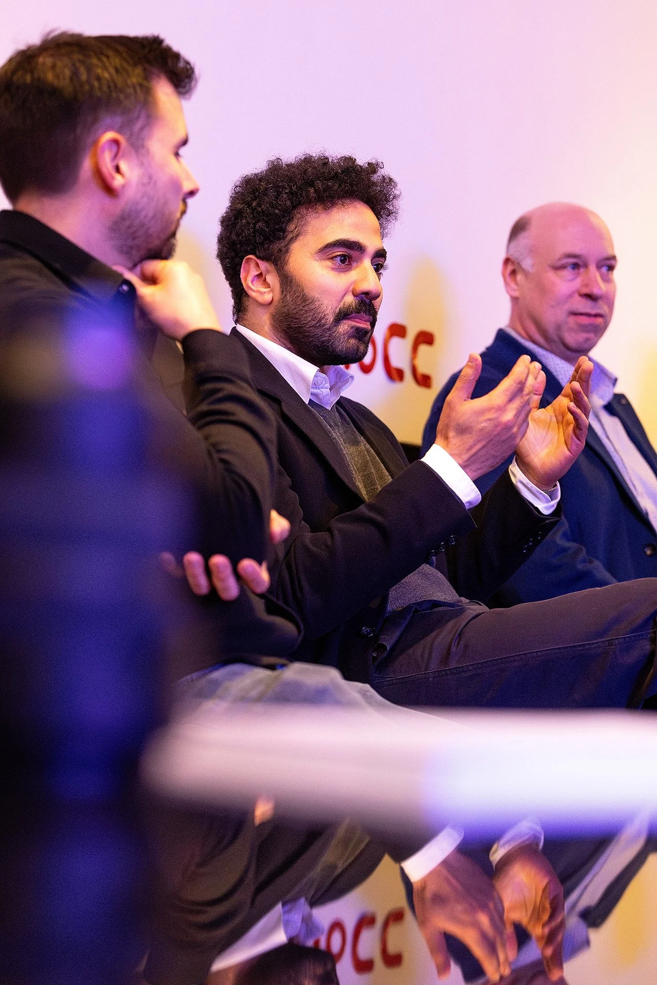 Three men seated in a row, engaged in a discussion or panel, with one man speaking or gesturing with his hand.