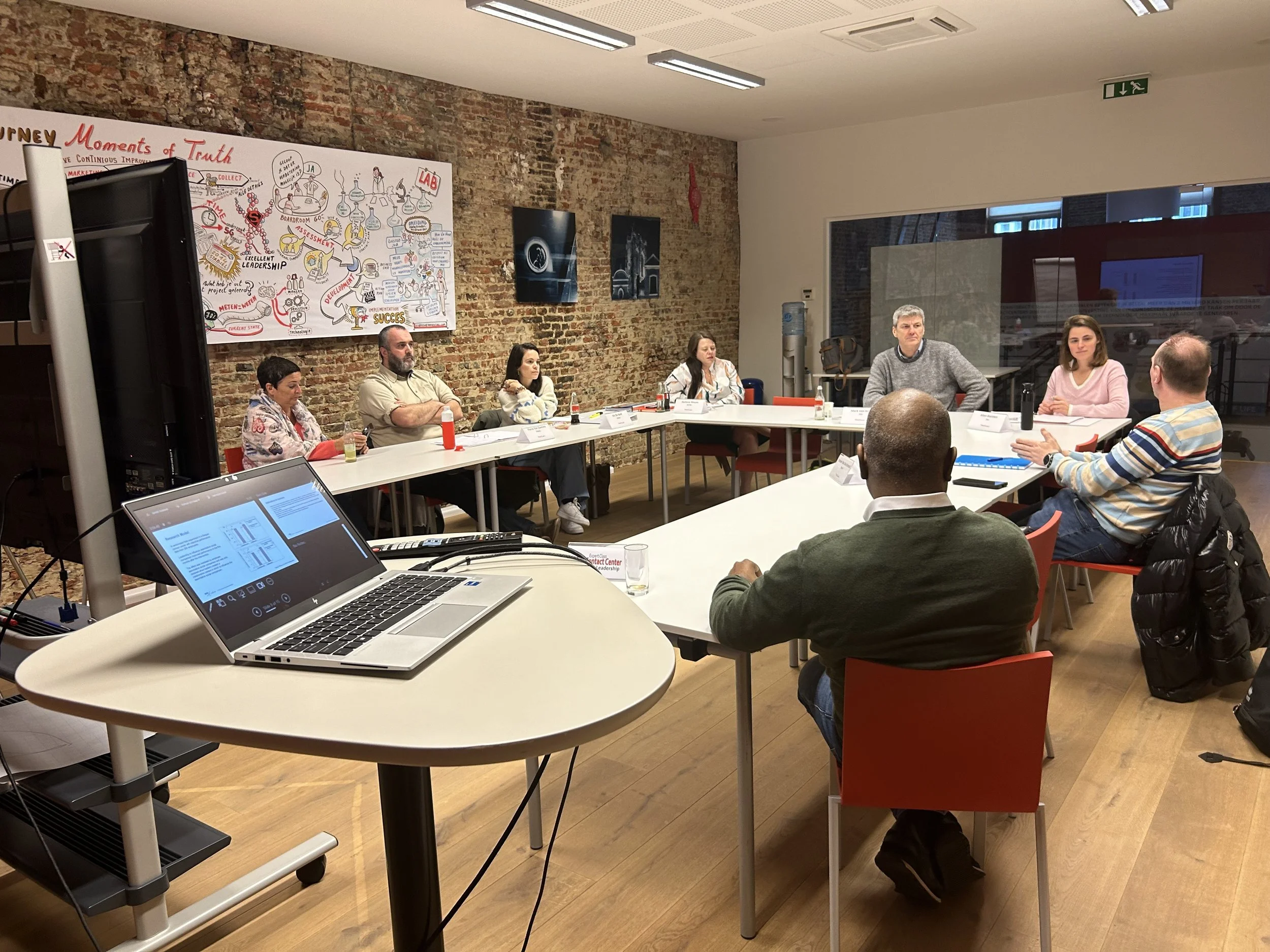 A group of people attending a meeting or workshop around a rectangular table in a modern conference room with brick walls. There are screens, papers, drinks, and a large illustrated poster on the wall.