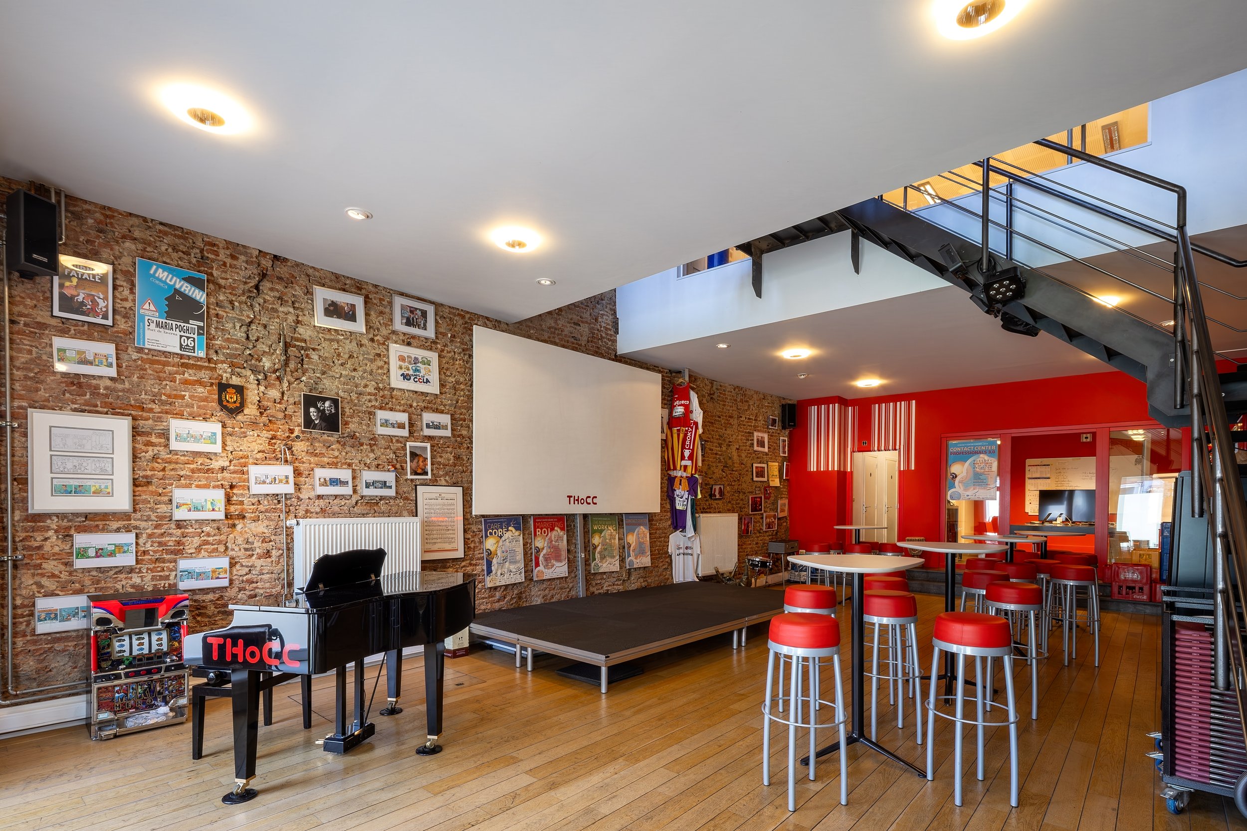 Indoor space with exposed brick walls and a mix of framed pictures and posters, a black grand piano, a small stage, and red and white barstools. There is a staircase with black metal railings leading upstairs, and bright ceiling lights.