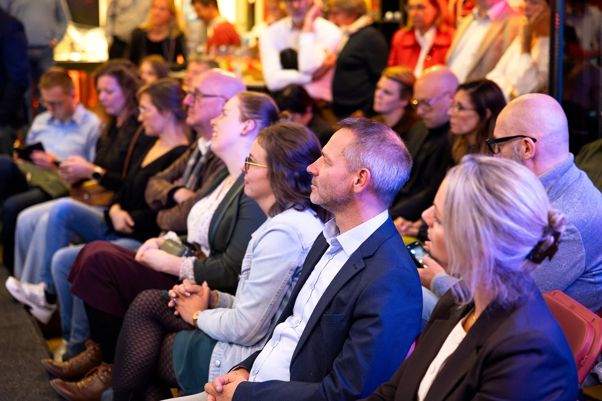 People seated in rows at a conference or event, paying attention to a speaker or presentation in a lively indoor setting.