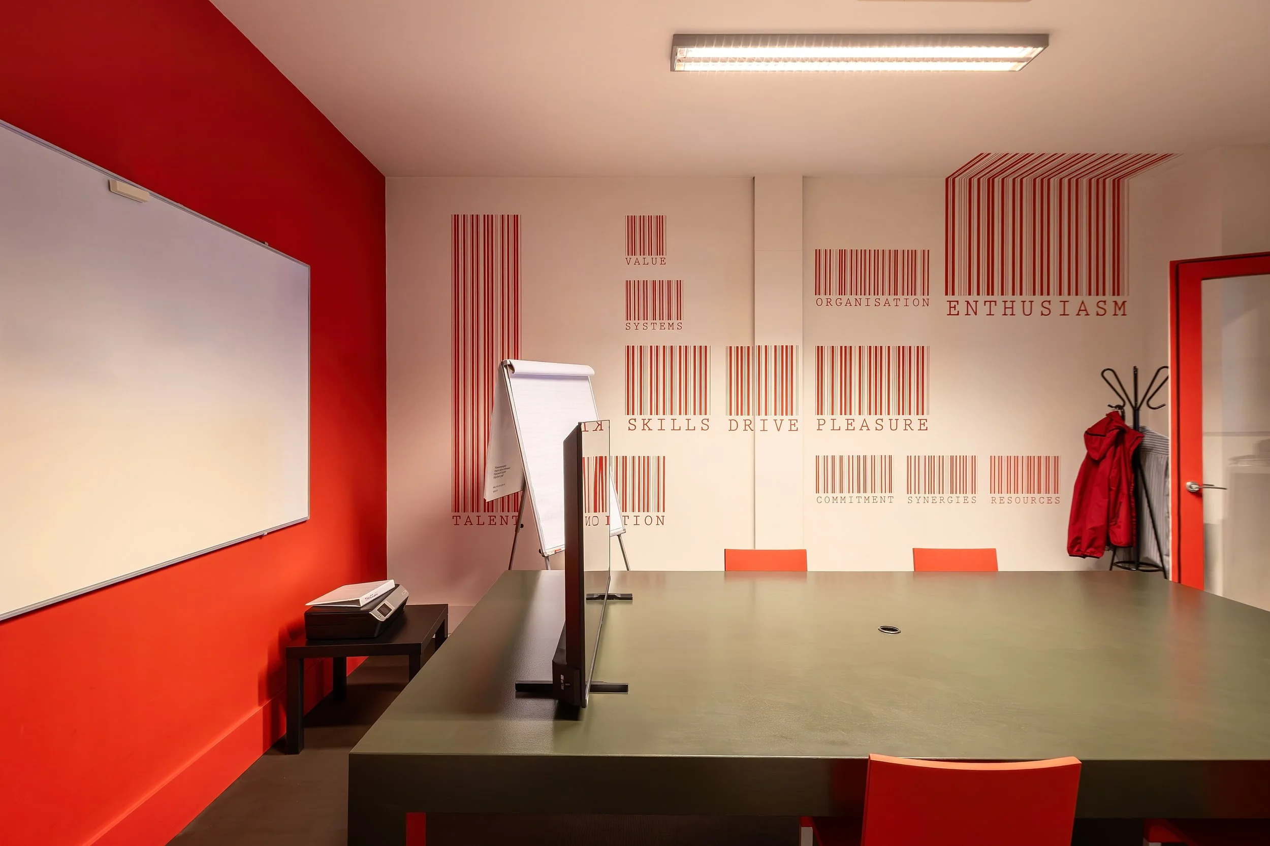 A modern conference room with a red and white color scheme, a large desk, a whiteboard, and wall art with words like 'value,' 'skills,' 'drive,' and 'pleasure' written alongside barcodes.