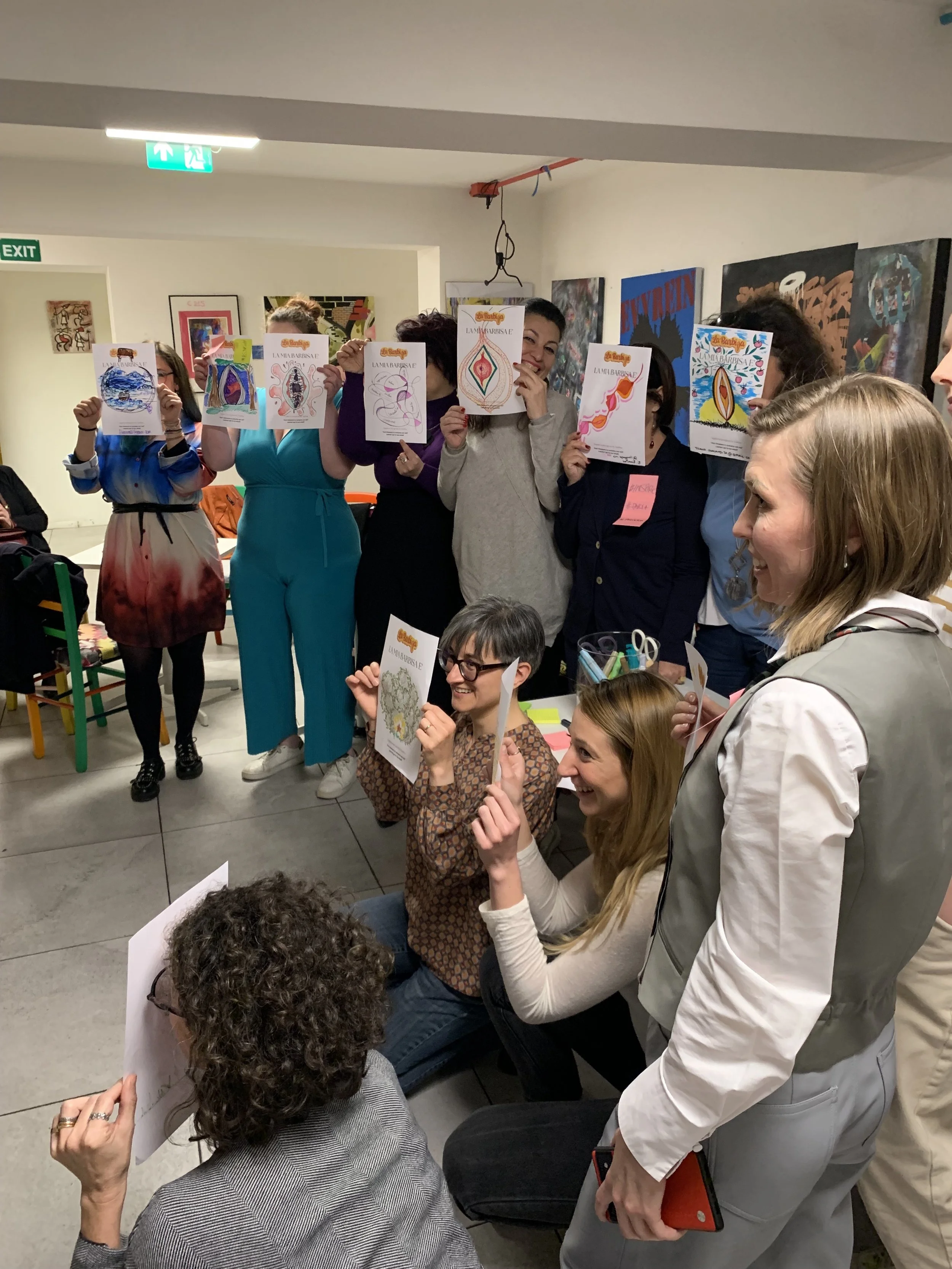 Gruppo di persone in una stanza che tengono dei fogli con disegni colorati davanti al viso, probabilmente durante un'attività artistica o workshop. Alcune persone sono in piedi e altre accovacciate, sorridono e mostrano i loro lavori.