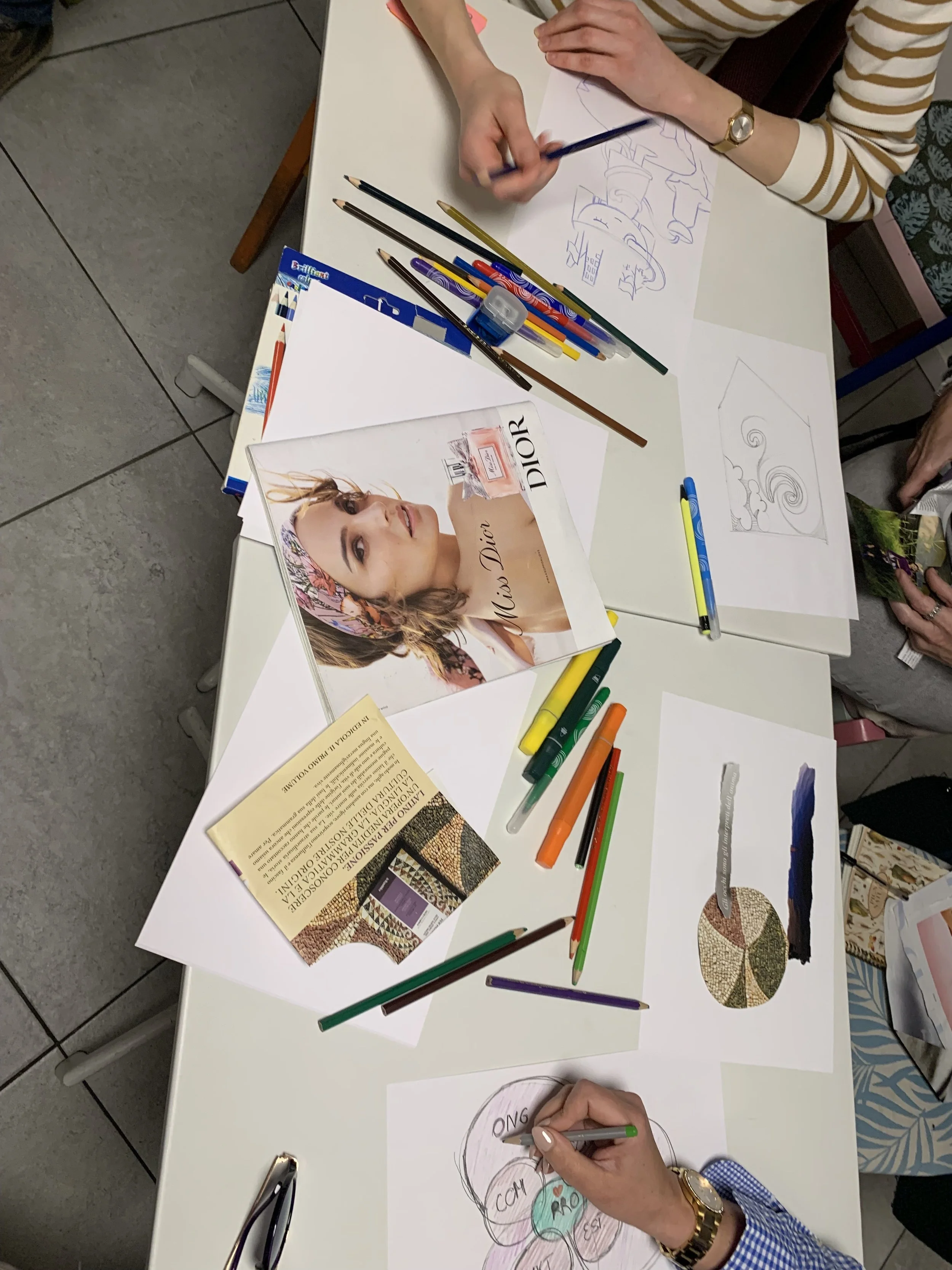 Persone che disegnano su un tavolo con fogli e materiale artistico, inclusi pennarelli colorati, matite e riviste. Una mano sta creando un disegno, altre mani sono visibili attorno al tavolo. Ci sono vari fogli con schizzi e colori.