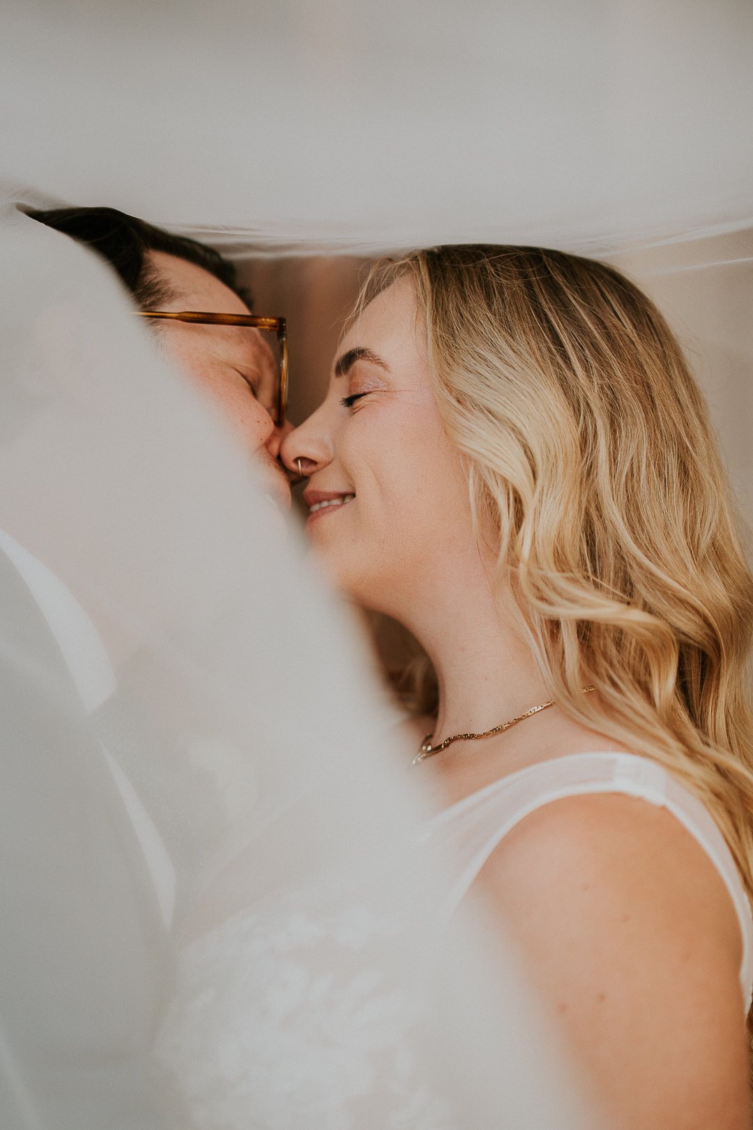 A couple in bed sharing a kiss, lying close together with eyes closed, smiling. The woman has blonde hair, a bracelet, and is wearing a white sleeveless top; the man has dark hair, glasses, and a nose ring.
