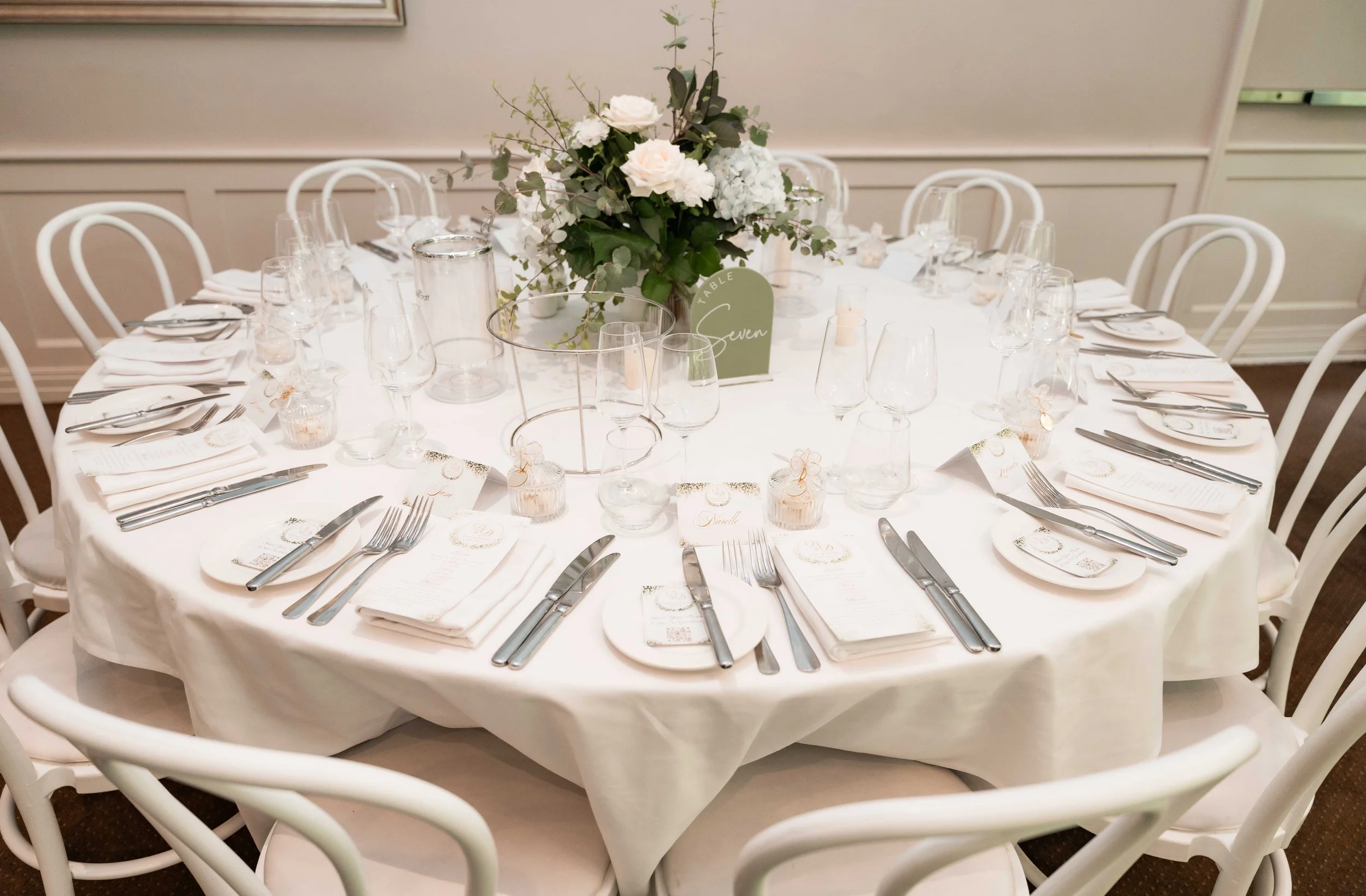 A round banquet table set for a formal event, with white tablecloth, neatly arranged silverware, glasses, napkins, and a centerpiece of white flowers and greenery. There is a small green table card indicating the table number seven.