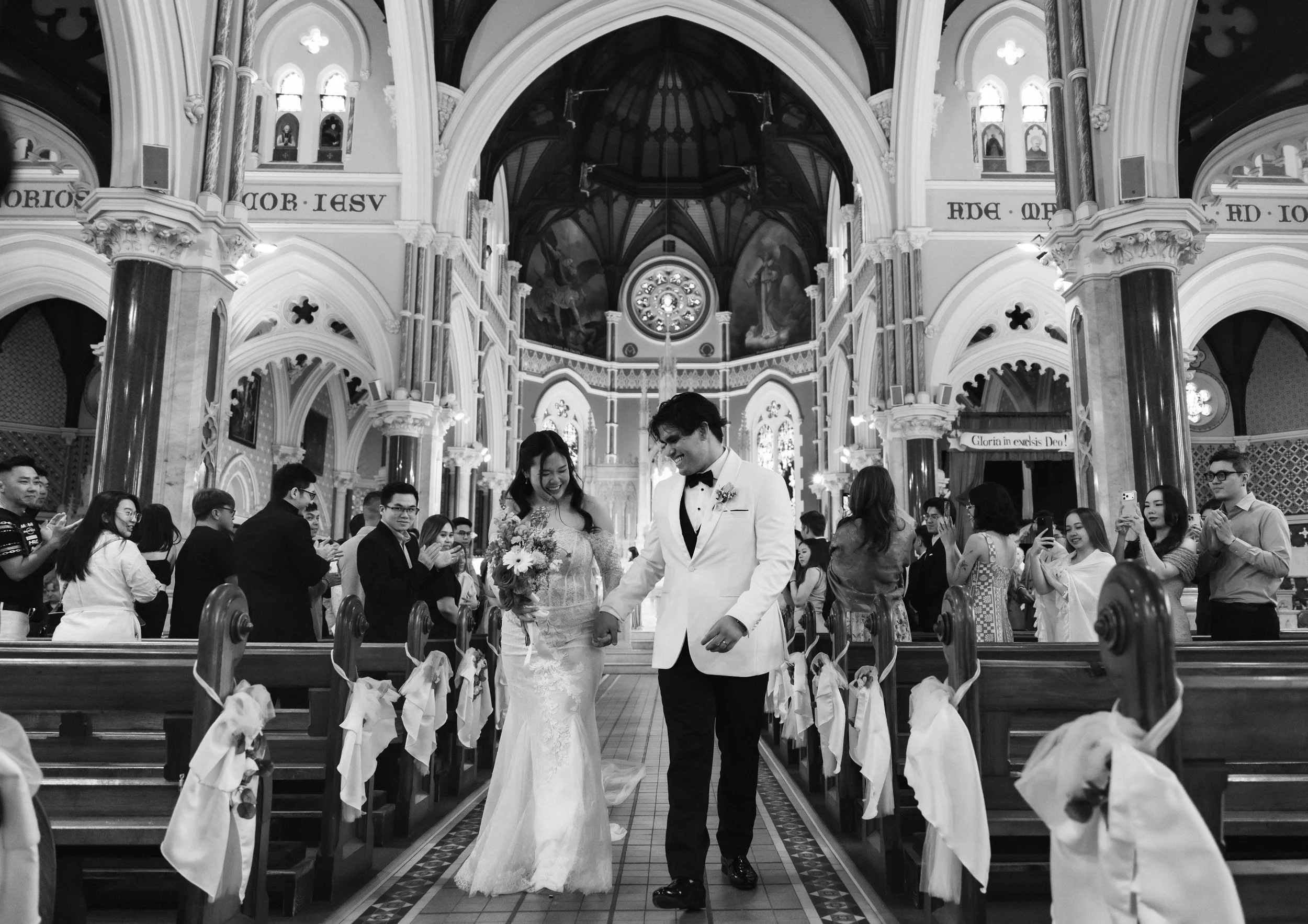 A bride and groom walking down the aisle of a church filled with guests during their wedding ceremony. The church has ornate architecture and stained glass windows.