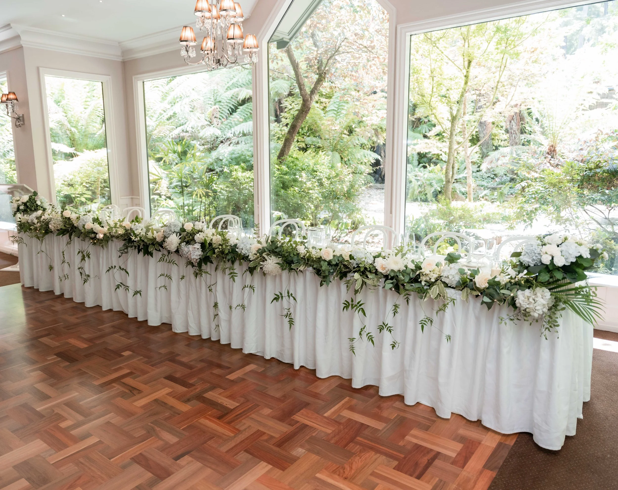 Wedding reception table setup with white tablecloth and floral arrangements, large windows showing trees outside, chandelier with lampshades hanging from ceiling.