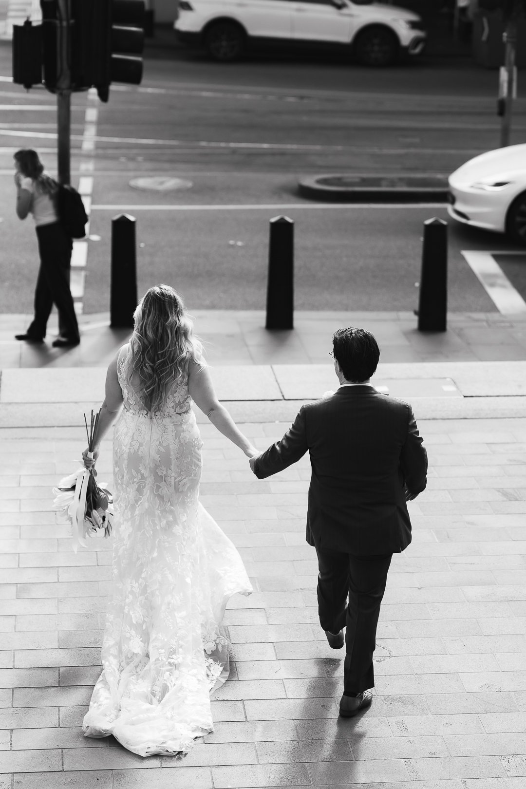 A bride and groom holding hands, walking up steps outside a building, with a city street in the background. The bride is in a lace wedding dress holding flowers, and the groom is in a dark suit.